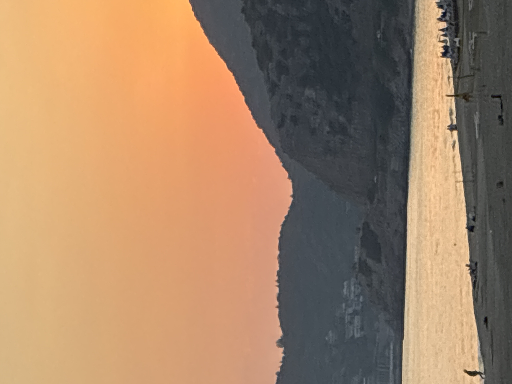 The image depicts a serene coastal scene at sunset in Alanya, Turkey. The sky is a gradient of warm orange and yellow hues, transitioning into a soft yellow near the horizon. In the foreground, a sandy beach is visible, with gentle waves lapping at the shore. A few people are scattered across the beach, some walking and others relaxing. Their forms are indistinct due to the distance and lighting.
Beyond the beach, a body of water, likely the Mediterranean Sea, reflects the warm colors of the sunset. The water appears calm and shimmering. In the background, rolling hills and mountains rise, their dark silhouettes contrasting with the bright sky. Some buildings are faintly visible on the slopes of these hills, indicating the presence of a settlement or resort. The overall atmosphere is tranquil and picturesque, characteristic of a Mediterranean evening.