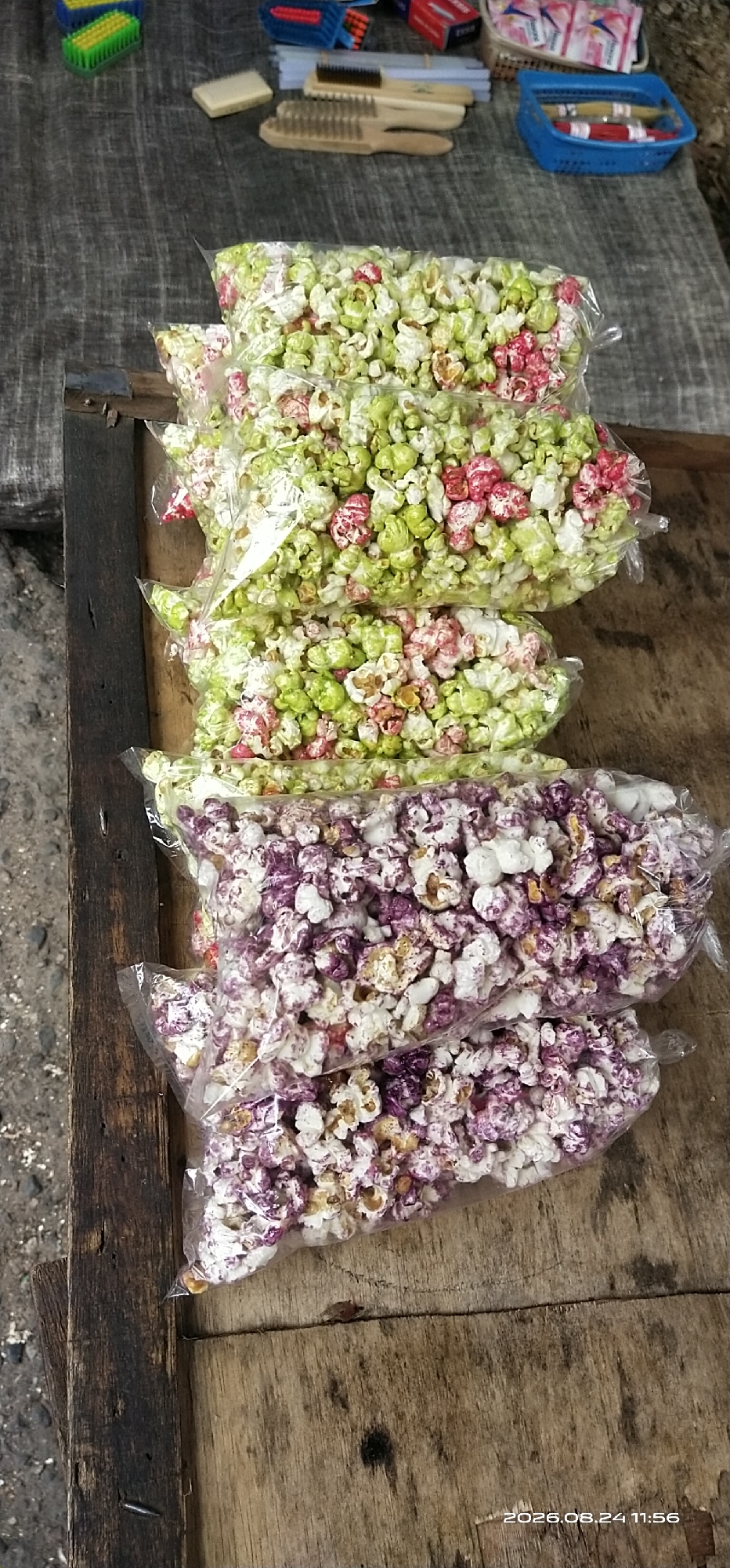 The image shows a close-up of several clear plastic bags filled with popcorn. The popcorn is multicolored, with varieties appearing to be green, pink, and white, and another variety appearing to be purple and white. The bags are stacked on a rustic wooden surface, which appears to be a table. In the background, various items are visible, including brushes with wooden handles, a blue plastic basket with what appears to be string or yarn, and a stack of small red boxes. The overall setting suggests an outdoor market or a vendor stall. A timestamp in the lower right corner of the image reads "2026.08.24 11:56", indicating the date and time the photo was taken. The location in Tamiang Layang, Indonesia, is not explicitly visible in the image but is stated as context.
