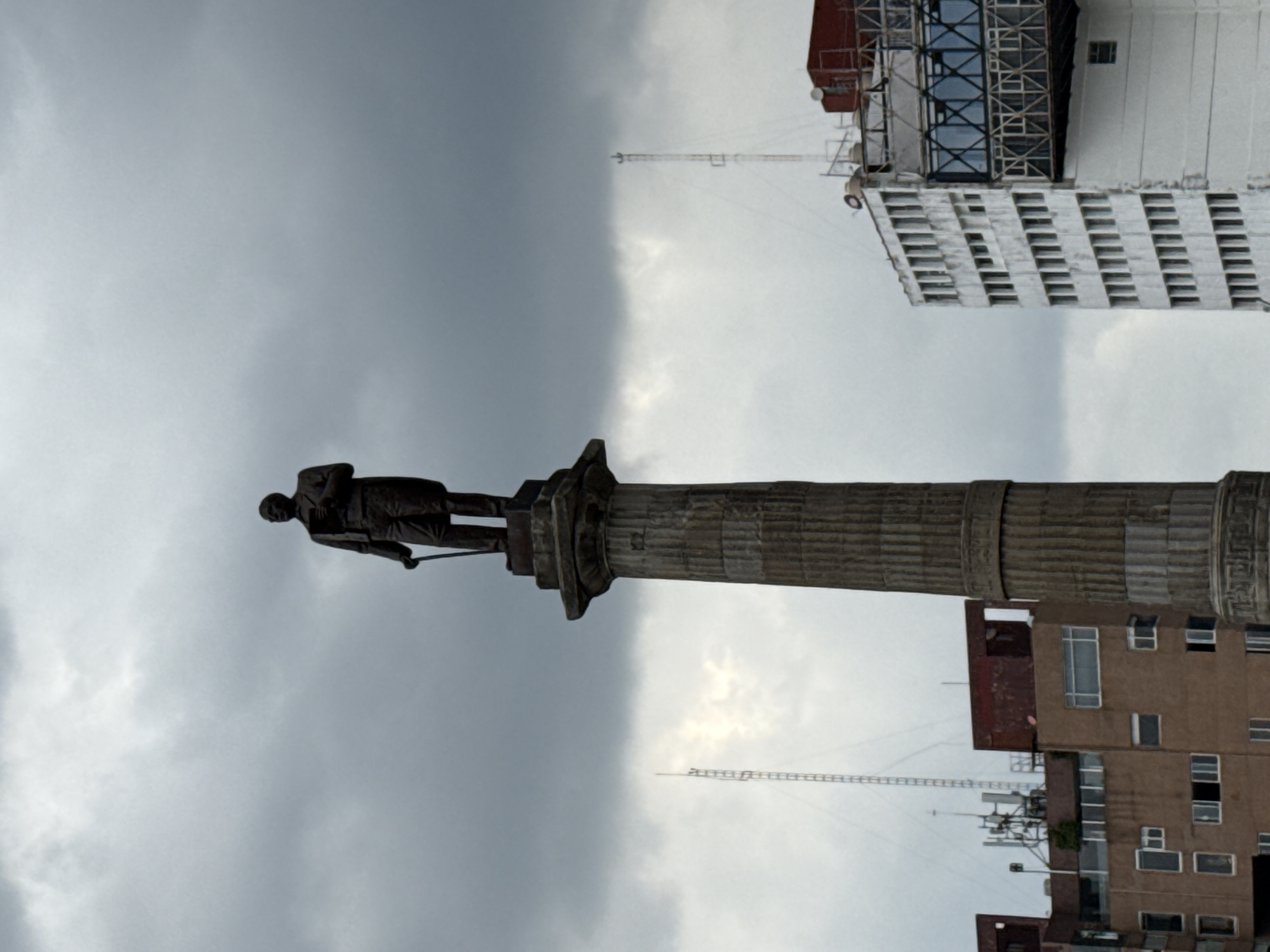 A statue of a man stands atop a fluted column in Puebla, Mexico. The statue is situated against a grey, cloudy sky. In the background, a white multi-story building with dark windows rises above other buildings. Multiple antennas are visible.