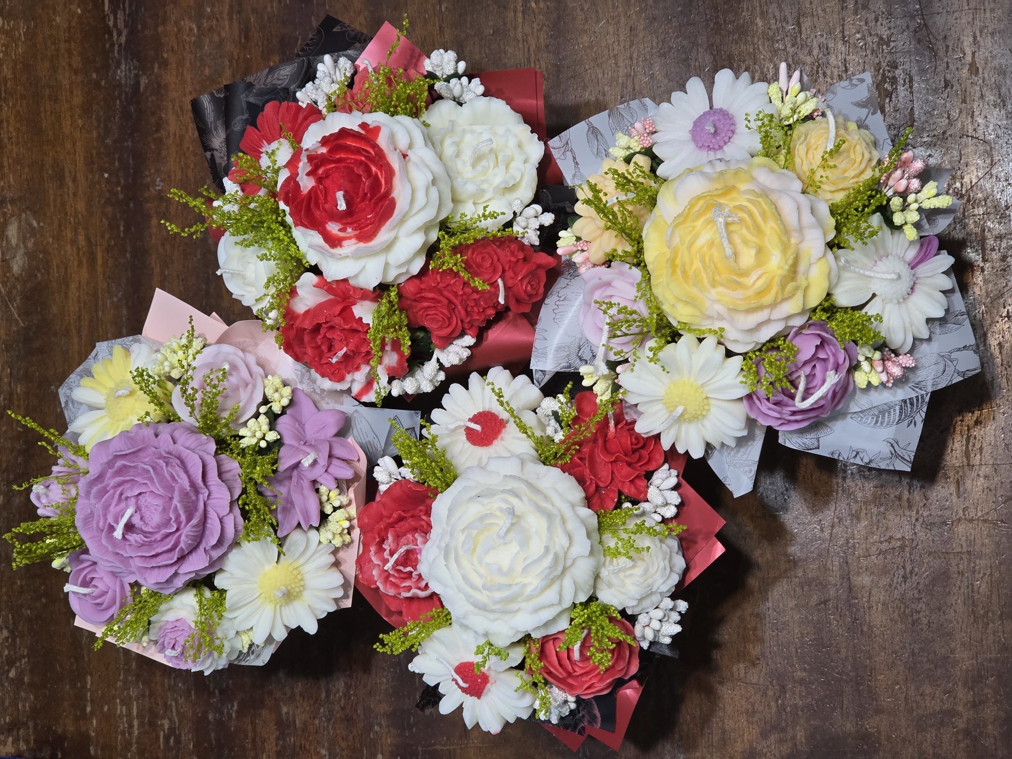 On a dark brown wooden surface are three bouquets of candle flowers wrapped in paper. The bouquet in the upper left contains white, red, and red-and-white candle roses arranged in black and red paper. The bouquet in the upper right has yellow and white candle flowers in grey patterned paper. The final bouquet in the lower left has purple and white candle flowers arranged in light pink paper. All bouquets are decorated with green sprigs and small white buds. The location is Unknown, Unknown.