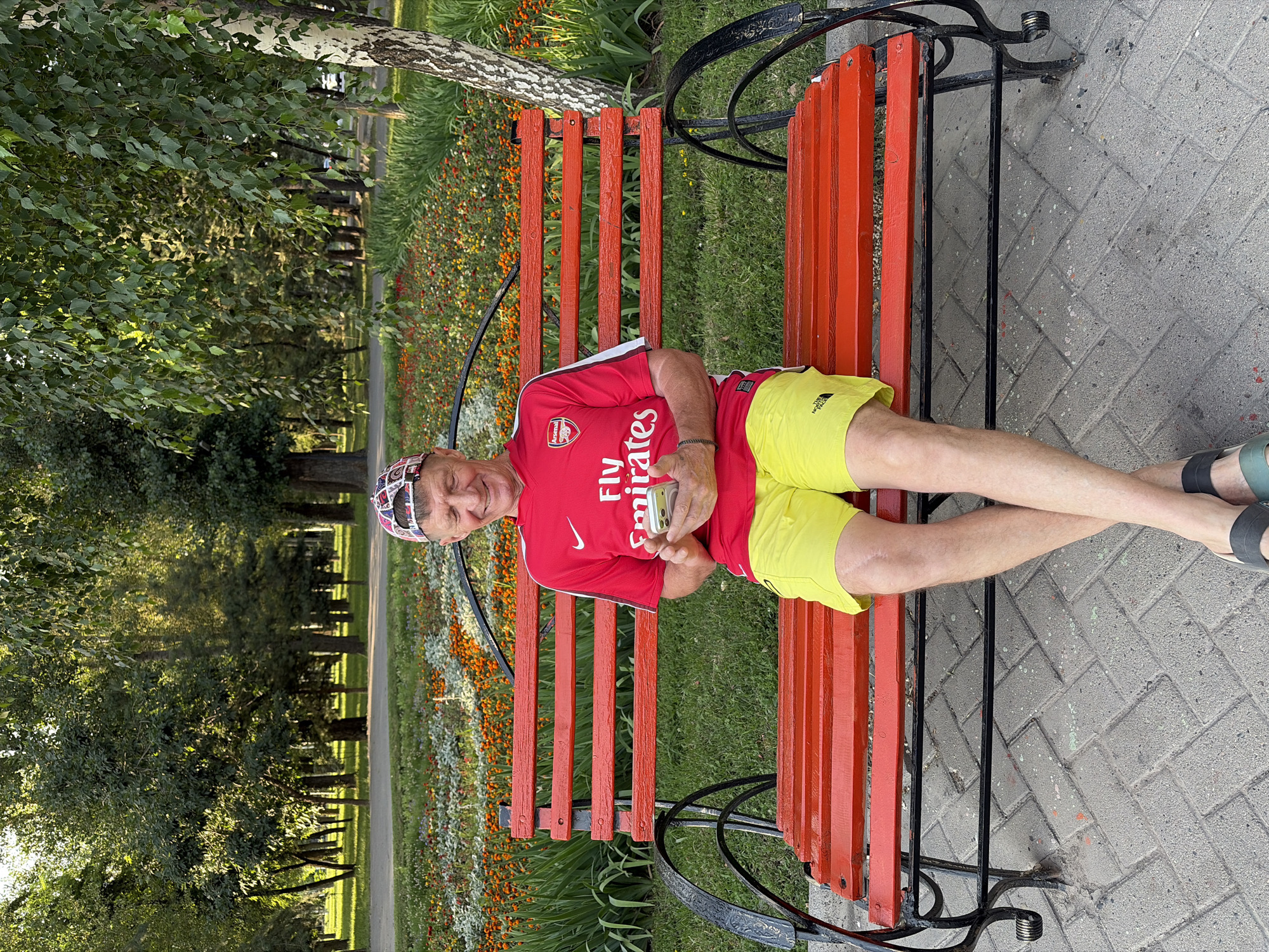 A man is sitting on a red park bench in a park. He is wearing a red Arsenal football jersey and yellow shorts, and a patterned baseball cap. He is looking at his phone and smiling. The bench is in front of a flower bed with orange and white flowers, and behind that are trees. The ground is paved with grey interlocking bricks.