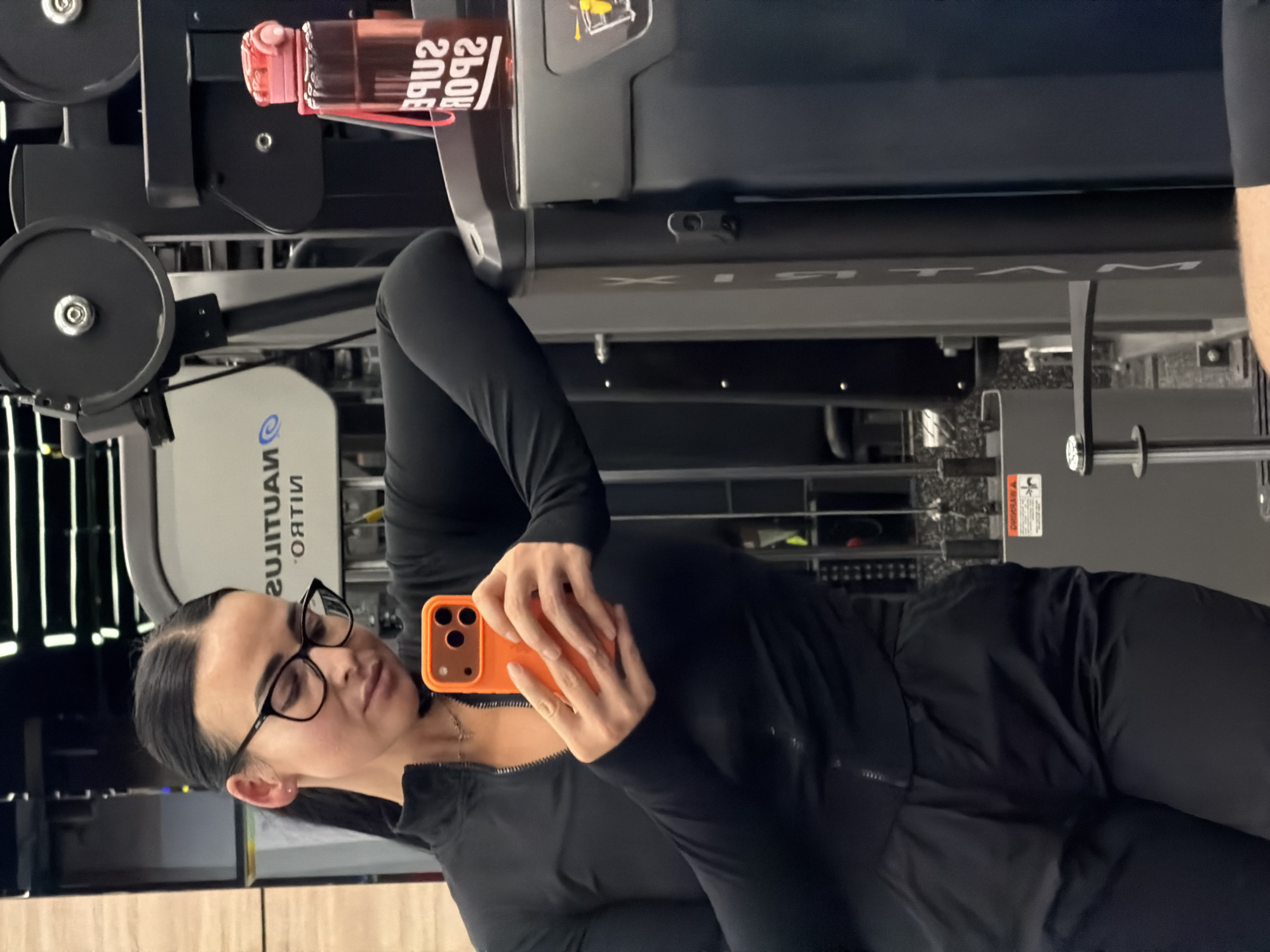 An image captures a woman taking a selfie in a gym, located in Saint Petersburg, Russia. She is depicted from the waist up, wearing a black, long-sleeved athletic top with a partial zipper and black pants. Her dark hair is pulled back, and she wears black-rimmed glasses. The woman holds a smartphone with a vibrant orange case in her right hand, angled to take a mirror selfie.
In the background, various pieces of gym equipment are visible. To her right, a pink or red water bottle, which appears to be hanging from a machine, displays the reversed text "SUPER SPORTS." Further back, gym machinery is seen, including one machine with the reversed brand name "NAUTILUS NITRO" and another with the word "MATRIX" prominently displayed. The setting suggests an indoor fitness center with typical gym lighting.