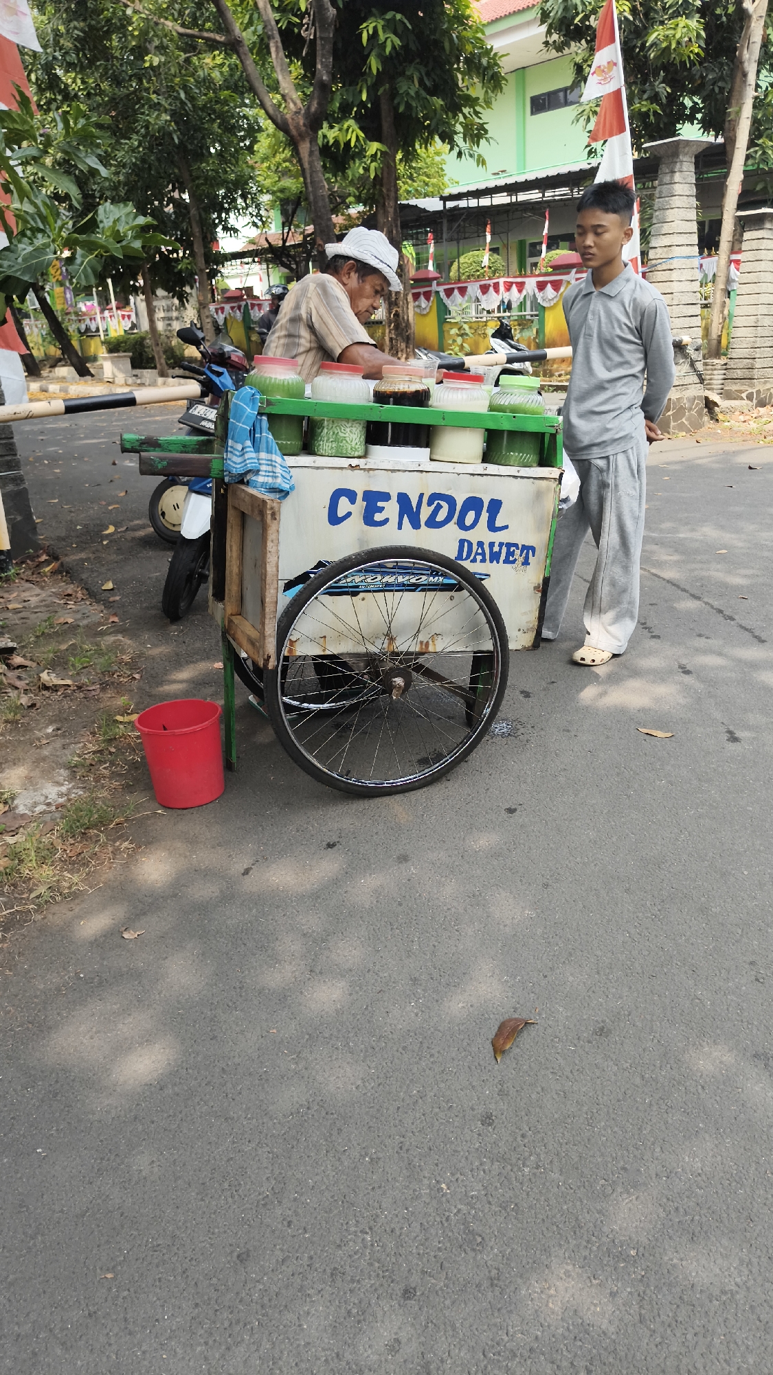 The image depicts a street scene in South Jakarta, Indonesia. A vendor, an elderly man wearing a white hat and striped shirt, is standing behind a cart selling cendol dawet, a traditional Indonesian dessert. The cart has a wooden frame and is painted white with blue lettering that reads "CENDOL DAWET". It contains several containers of colorful ingredients. To the right of the cart, a young man in a grey long-sleeved shirt and grey pants stands with his hands behind his back, appearing to be a customer or assistant. A red bucket is visible to the left of the cart, and a motorcycle is parked behind it. The background shows trees, a building with a green roof, and a small Indonesian flag. The scene is set on an asphalt road with some scattered leaves.