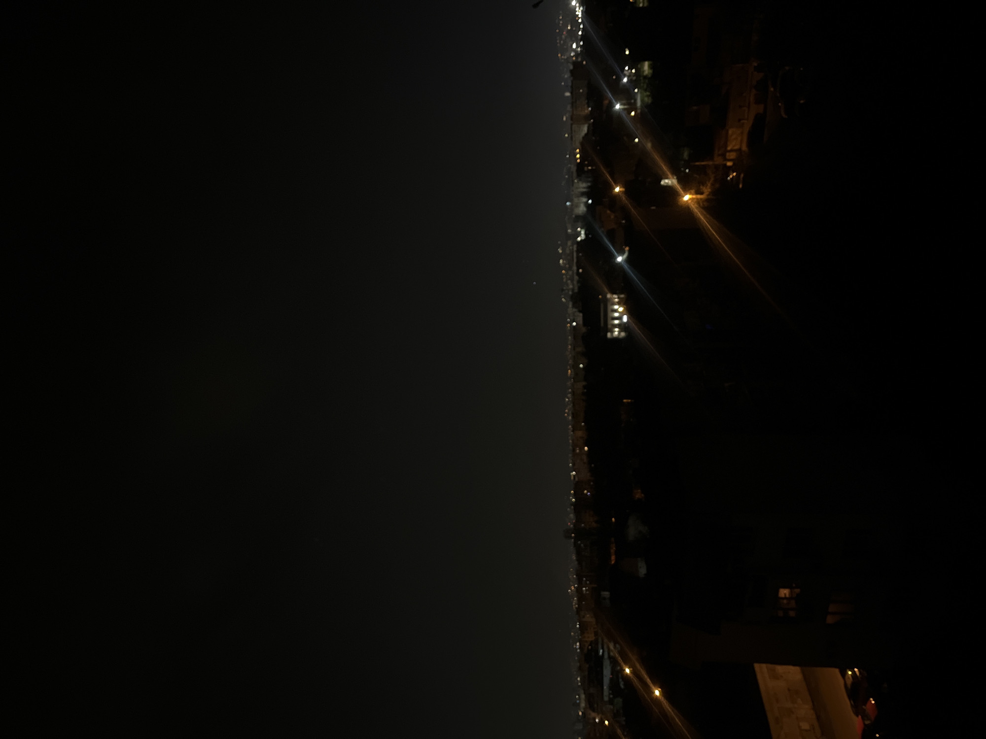 The image shows a nighttime view of what appears to be a coastline or cityscape with a large body of water or dark expanse on the left side. A line of lights runs vertically, likely representing a road, streetlights, or buildings along the shore. The right side of the image is mostly dark, with sparse lights suggesting buildings and structures. There are no discernible people, actions, or interactions directly visible. Given the prompt's reference to Sofia, Bulgaria, the water body might be a large lake or reservoir near the city, though this cannot be definitively confirmed from the image alone. More context would be required to confirm the specific location.