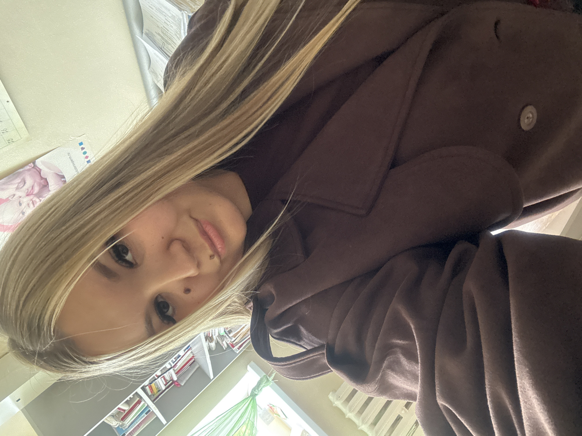 A young woman with blonde hair is looking down at the camera. She has fair skin and a mole on her face. She is wearing a brown coat. Behind her is a bookshelf with books and a window. The location appears to be indoors, likely a home in Фоки, Russia, given the city context provided.