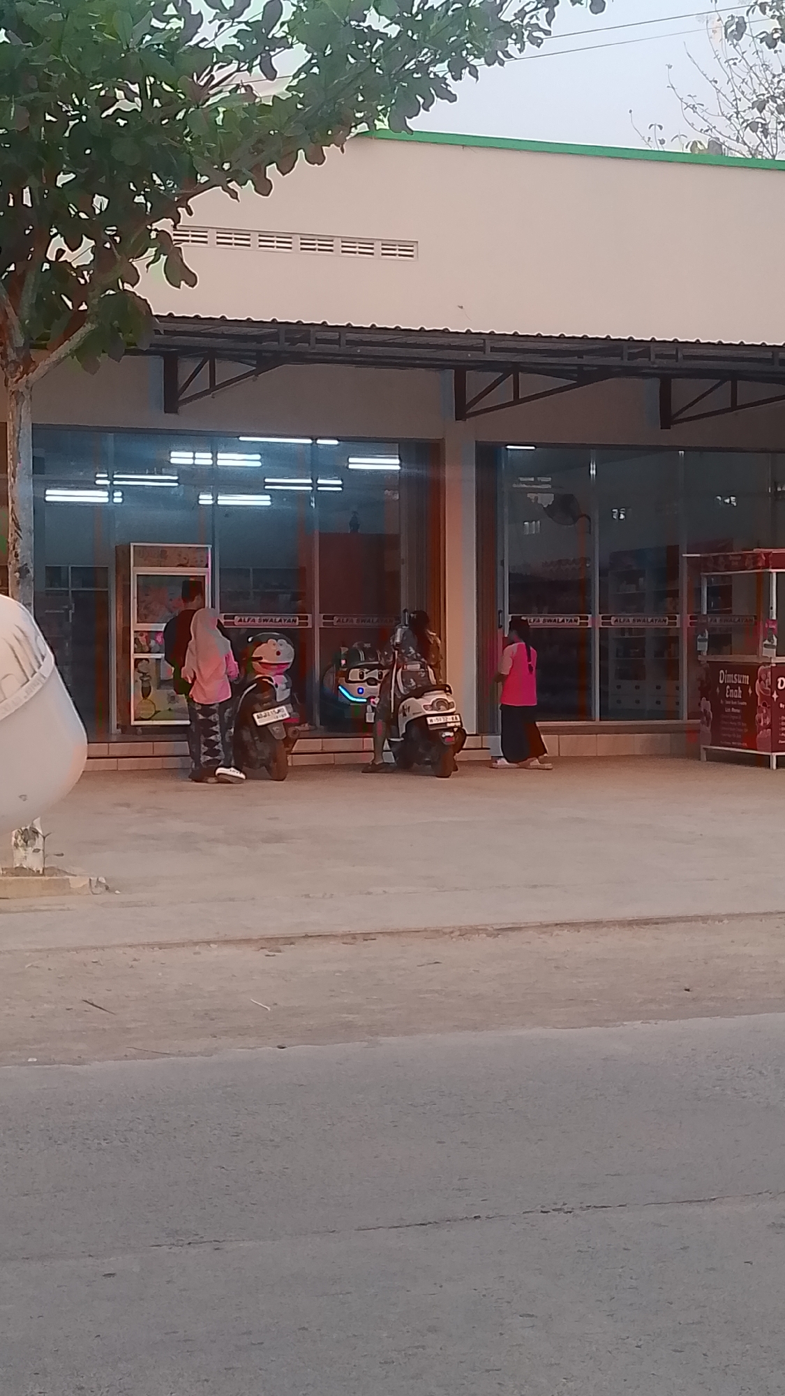 The image depicts an outdoor scene in Karanglangu, Indonesia, featuring a storefront with large glass windows. In front of the store, there are two motorcycles parked on a paved surface. Three individuals are visible. One person, wearing a pink headscarf and a patterned outfit, stands near a motorcycle. Another person, dressed in a black shirt and pants, stands beside them. A third individual, wearing a pink top and dark pants, is seen walking away from the storefront. The storefront itself has a white facade with a green trim along the top. Signs with text, including "ALFA SWALAYAN," are visible above the glass windows. Part of a tree with green leaves is present on the left side of the frame. The background suggests a moderately lit environment.