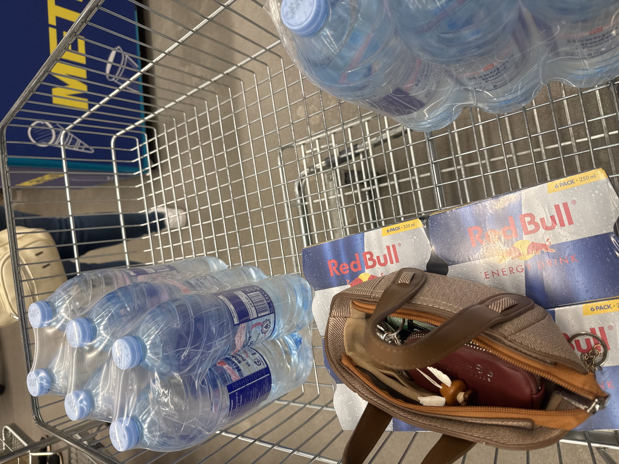 An indoor, high-angle shot captures the contents of a metal wire shopping cart, suggesting a shopping trip. The cart contains several items, including multiple packs of bottled water and two 6-packs of Red Bull energy drinks.
On the left side of the cart, six large plastic water bottles are wrapped together, with light blue caps. Cyrillic text is visible on their labels, including "БАЛДАРАН" (Baldaran), "Извор" (Izvor - Spring), "Родоп" (Rodop), and "1,5 L" indicating the bottle volume. Another pack of water bottles is seen in the upper right.
Towards the right, two multipacks of Red Bull energy drink are stacked. The packaging clearly shows the "Red Bull" logo, the words "ENERGY DRINK", and "6 PACK • 250 ml". A small, open patterned purse or handbag with brown leather straps rests on top of one of the Red Bull packs. Inside the purse, a set of keys with a distinct mushroom-shaped charm and a small red wallet are visible.
In the background, behind the shopping cart's wire frame, a blue surface with large yellow letters spelling "IKEA" is partially visible, indicating the location is likely an IKEA store. The scene appears to be well-lit, typical of a retail environment. No people are visible, and no specific time of day or emotional state can be inferred.