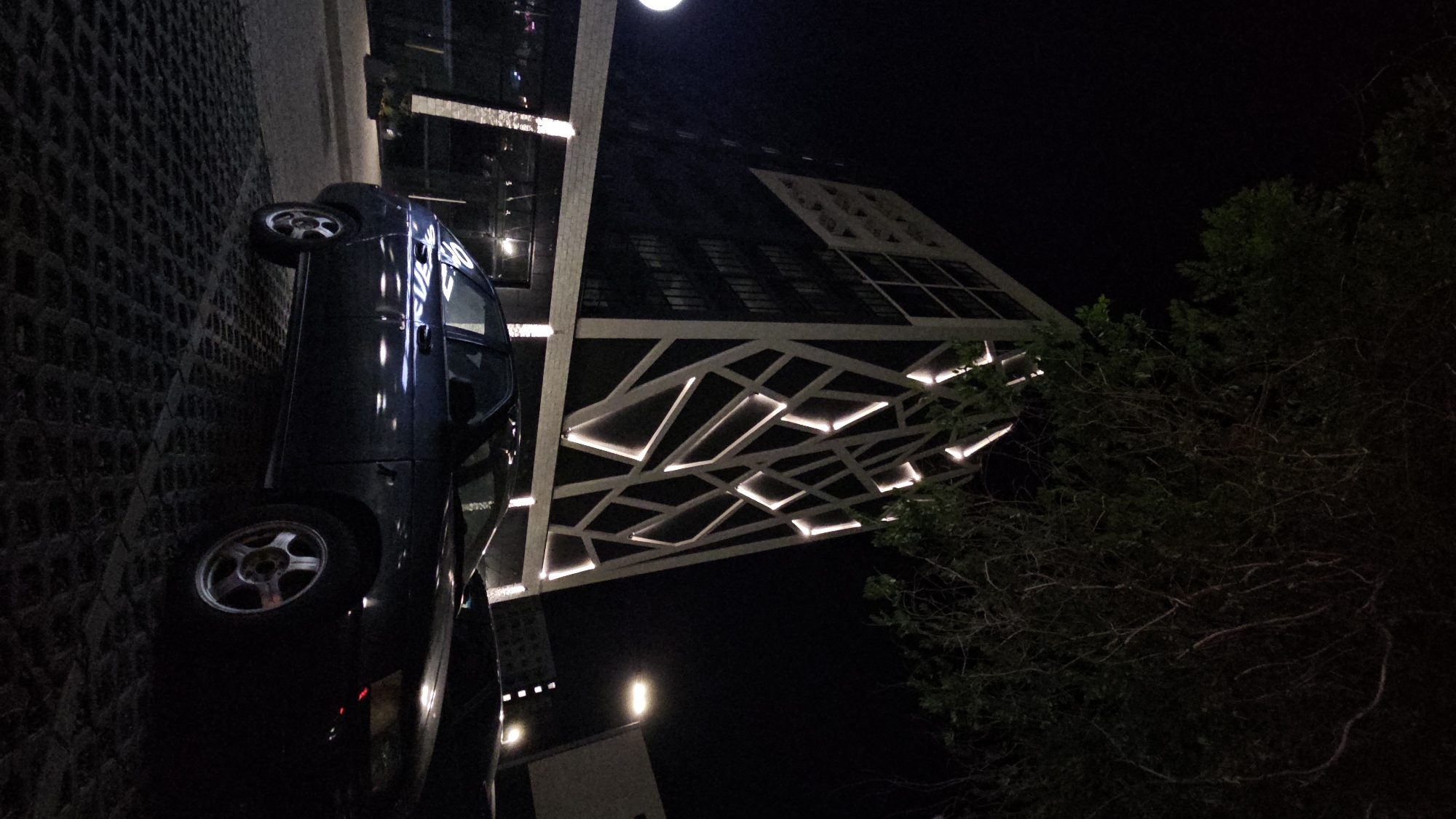 The image shows a dark, possibly nighttime scene in a city, with a modern building illuminated by embedded lights. The building features a distinctive geometric facade with white lines forming a pattern that appears to be integrated with lighting elements, creating a striking visual effect against the dark structure. In the foreground, a dark-colored sedan is parked on a textured surface, likely a street or parking lot. The car is positioned at an angle, showing its side and front. Above and to the left, a large tree with dark foliage partially obscures the sky and extends over the scene. Streetlights are visible in the background, contributing to the urban ambiance. Based on metadata, this image was taken in Ulyanovsk, Russia.
