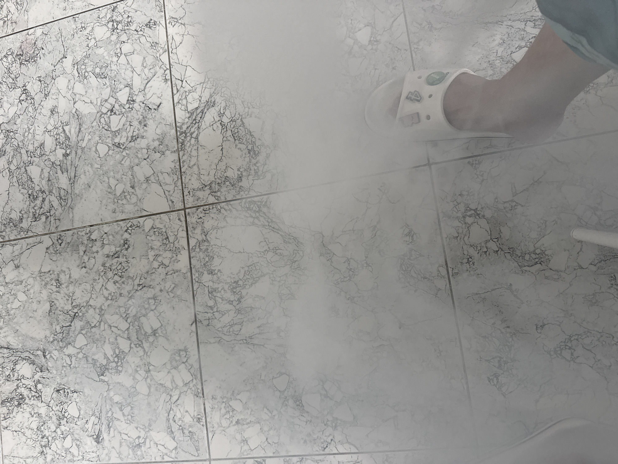 The image shows a close-up of a tiled floor with a marble-like pattern. A person's foot, wearing a white slip-on sandal with several small decorative charms, is visible on the right side of the frame. The scene is partially obscured by a hazy white mist or steam, which creates a diffused lighting effect and obscures details in the background. The tiling appears to be in a domestic or commercial setting, potentially a bathroom or spa, given the presence of mist. The location is Yakutsk, Russia.