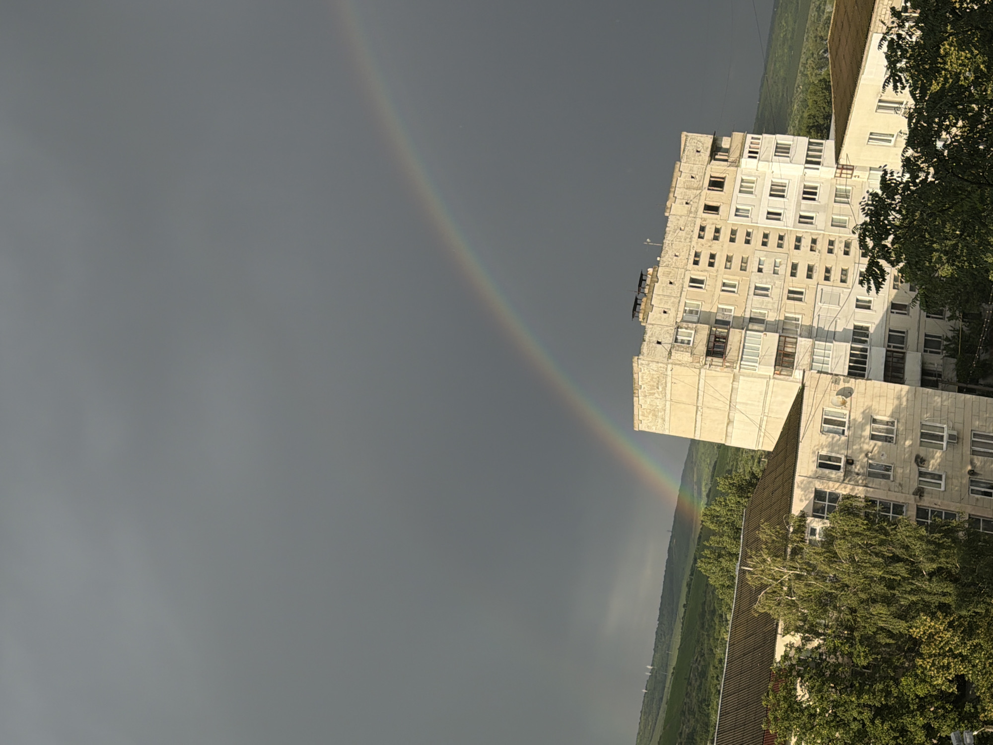 A vertical, slightly tilted shot captures a segment of a city in Strășeni, Moldova, under a dark, overcast sky. A faint rainbow arcs across the upper portion of the frame, its colors muted against the somber clouds. In the midground, a tall, pale apartment building dominates the scene, its facade weathered and showing signs of age. Numerous windows, some with balconies, punctuate its structure. Below and to the left of this building, a lower, darker-roofed building is partially visible, also featuring windows and what appears to be a satellite dish. Lush green trees, some quite tall and dense, are interspersed between the buildings and in the foreground, their leaves vibrant against the muted tones of the architecture and sky. In the background, rolling green hills rise, their slopes dotted with more trees, suggesting a semi-urban or rural fringe location. A few distant, slender structures, possibly utility poles or small towers, are visible on the hilltop. The overall impression is one of an ordinary urban landscape momentarily enhanced by the ephemeral beauty of a rainbow.