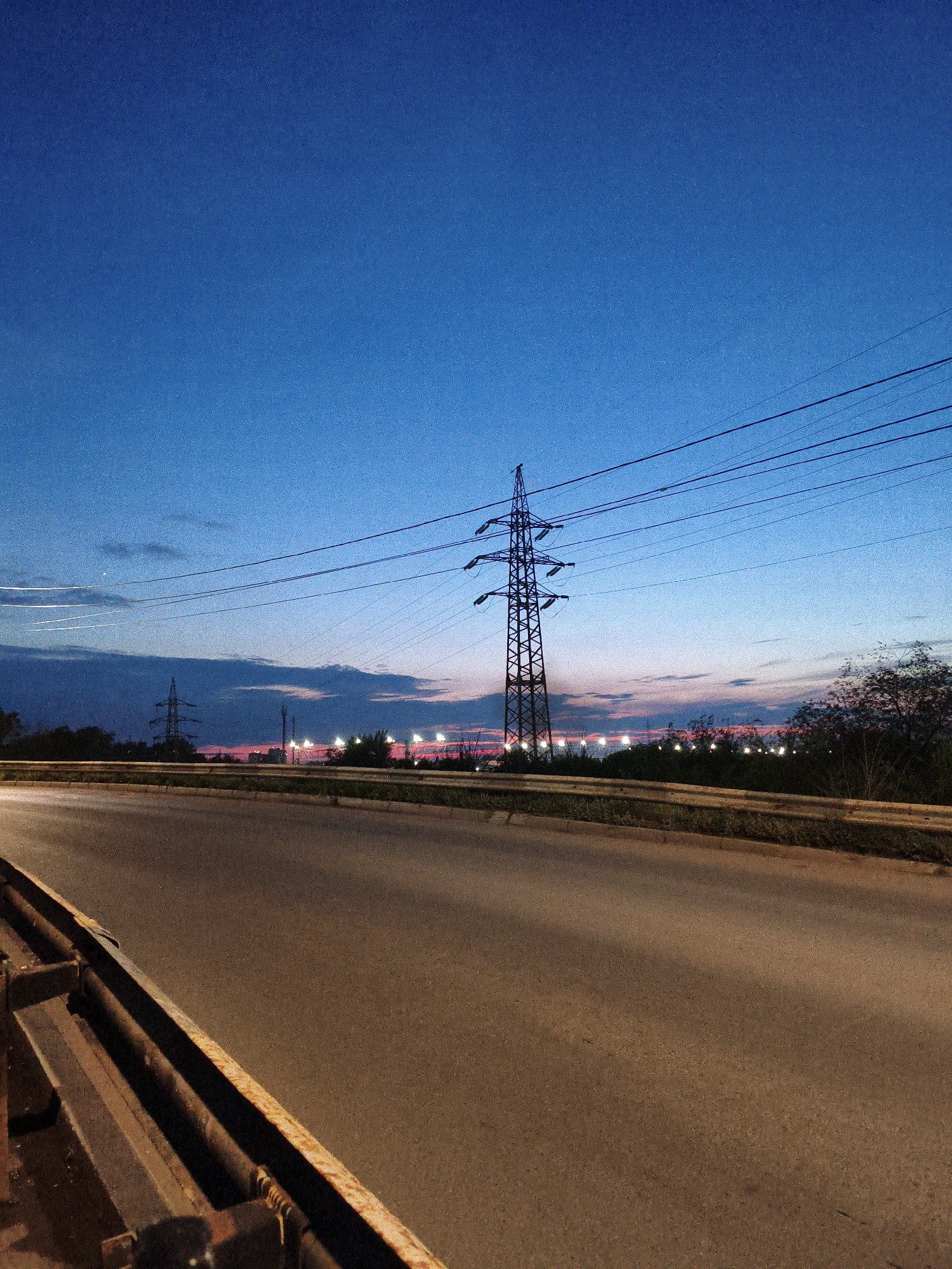 The image captures a twilight scene on a road in Orenburg, Russia. A highway stretches into the distance, bordered by a rusted metal guardrail on the left and a grassy verge on the right. In the background, a tall electricity pylon stands prominently against a vibrant blue and pink sky, indicating the transition from day to night. Several streetlights are visible along the horizon, and distant trees form a dark silhouette. The sky shows faint clouds and light streaks, possibly from passing aircraft. The road surface is asphalt, reflecting the ambient light and showing tire marks. There are no people or animals visible in the frame.