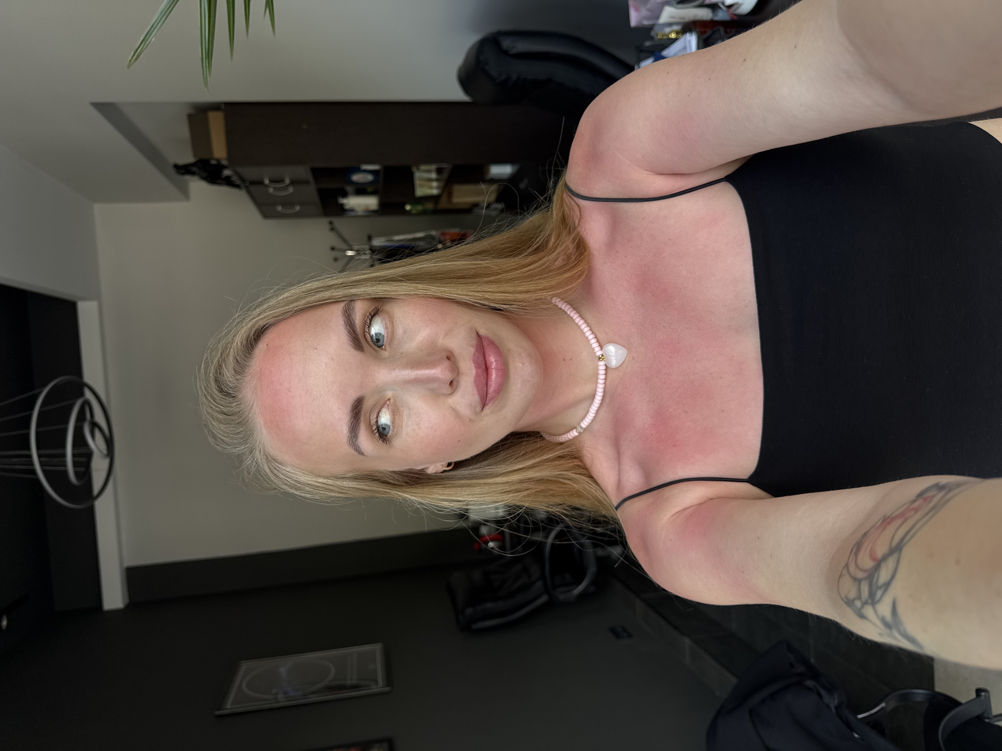 A woman with blonde hair and fair skin is centered in the image, wearing a black tank top and a pink beaded necklace with a white heart pendant. She is taking a selfie, holding the camera at arm's length, and her arm has a tattoo visible on it. The woman appears to have some sun exposure on her face and shoulders. Behind her is a dimly lit room with dark walls and a dark brown cabinet. A decorative metal object is visible on the left, and a framed picture is on the wall. There is also a plant visible in the upper left corner of the frame. It is not possible to determine the location in the city of Sochi, Russia, based on the image provided.