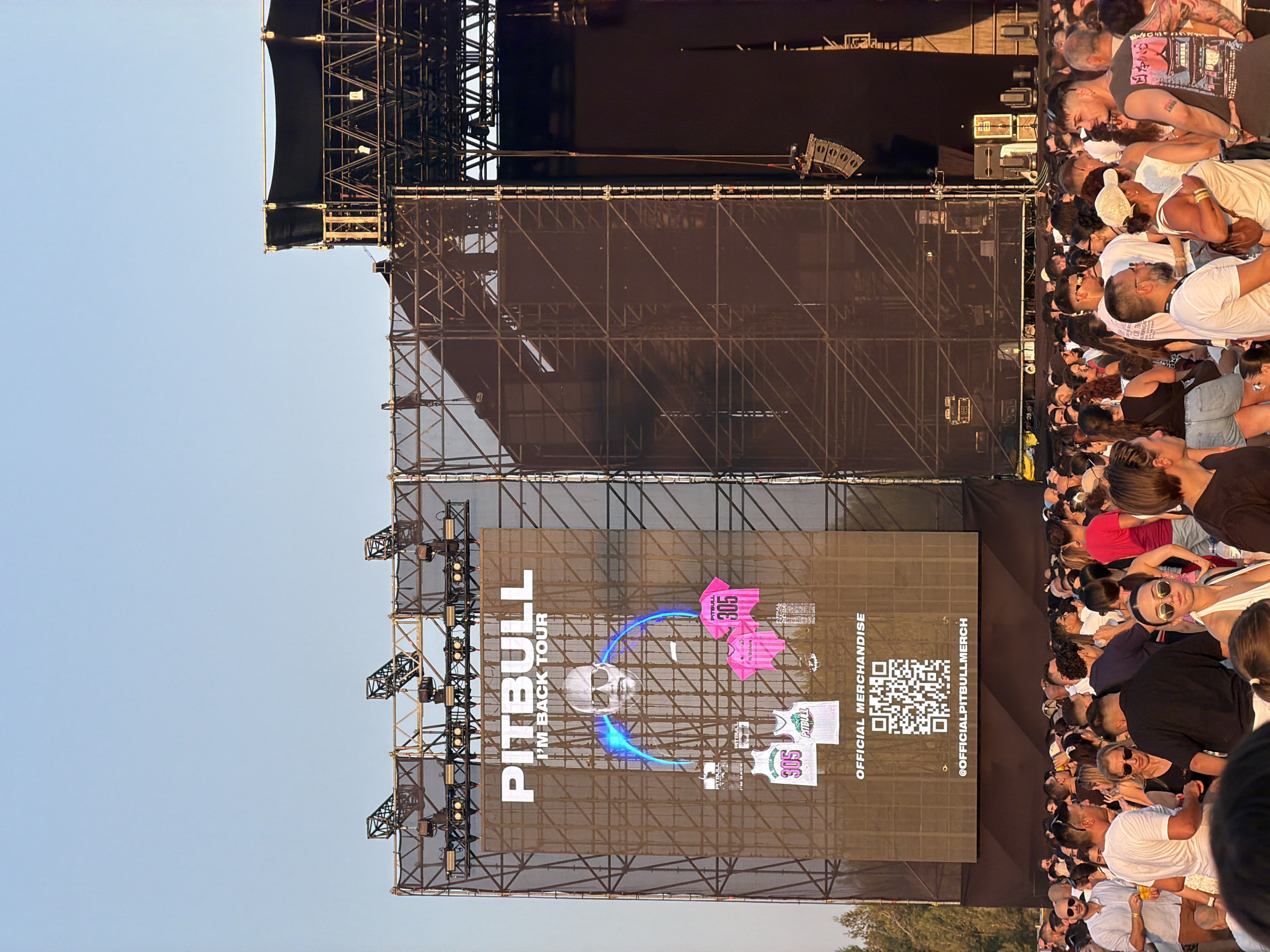 The image captures an outdoor concert scene, likely in Mosta, Malta, during daylight. Dominating the frame is a large stage structure with a massive screen displaying a promotional image for the "PITBULL I'M BACK TOUR." The screen features a portrait of Pitbull, along with graphics of merchandise like jerseys and t-shirts, and a QR code for official merchandise. A large crowd of people is gathered in front of the stage, suggesting they are attending the concert. The environment appears to be an open area, possibly a large field or plaza, with some trees visible in the background. The overall lighting indicates it is daytime, with the sun casting shadows and illuminating the scene.