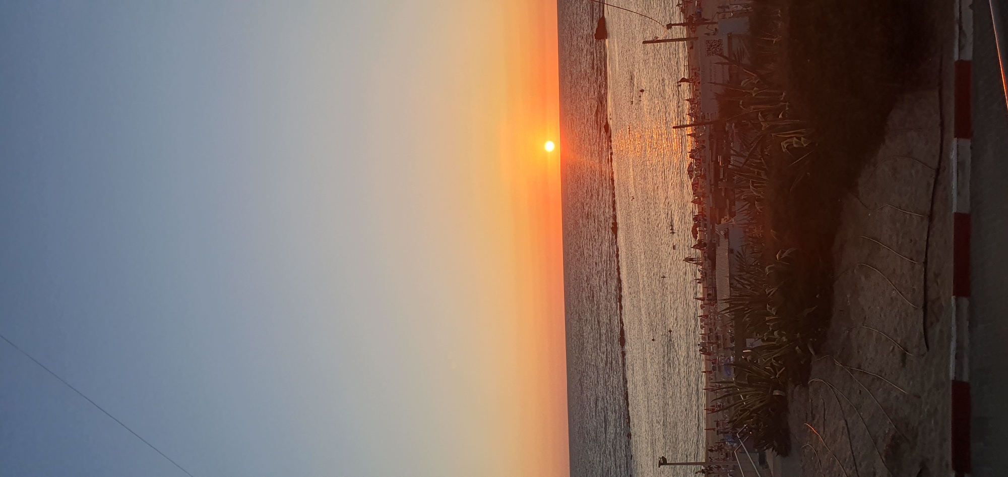 The image captures a sunset scene at a beach in Bat Yam, Israel. The sun, a bright orb, is setting over the calm, shimmering sea, casting a warm, golden-orange glow across the sky and reflecting on the water's surface. The sky transitions from a pale blue at the top to a vibrant orange near the horizon.
The beach is active with people enjoying the evening. Umbrellas and lounge chairs are set up, suggesting a popular tourist or recreational spot. Some individuals are visible in the water, while others are walking along the shoreline or relaxing on the sand. Lush green plants, possibly palm trees, are scattered along the edge of the beach, adding to the tropical ambiance.
In the foreground, a sandy path is visible, marked by a red and white striped barrier along the right edge, possibly indicating a walkway or the boundary of the beach area. The overall mood is tranquil and serene, with the warm colors of the sunset evoking a sense of peace and relaxation. The time of day is clearly sunset, and the weather appears to be clear and pleasant. There is some visible text on a white structure on the beach, which is difficult to read clearly but appears to include a QR code.