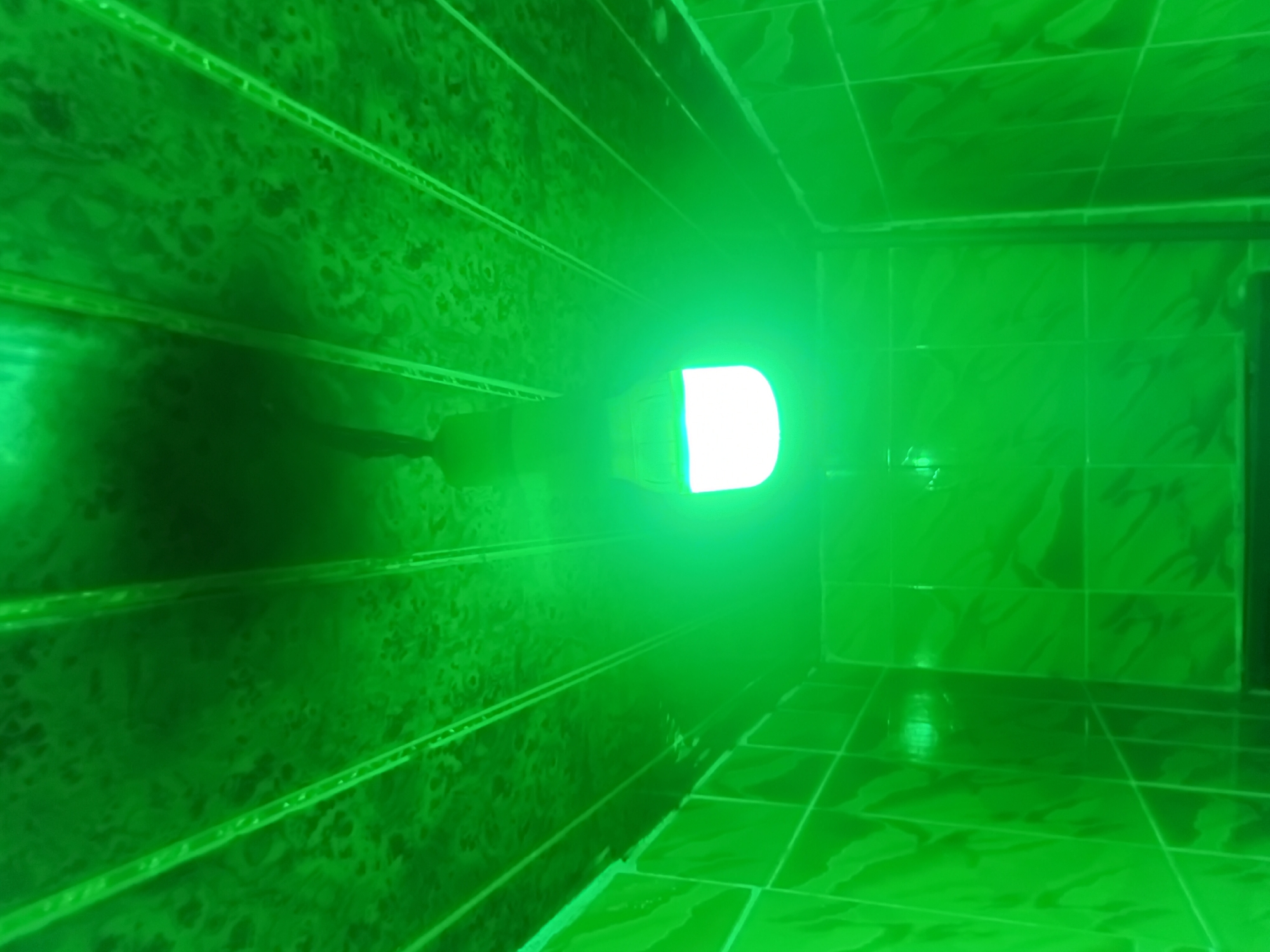 The image shows a bathroom with green tiles and a green light bulb hanging from the ceiling. The ceiling also appears to be covered in green material, possibly tiles or a paneling. The walls and ceiling are illuminated by the green light bulb, creating a predominantly green environment. The lighting is quite bright, with some areas of the ceiling showing shadows and texture. The tiles on the walls are square and have a patterned surface. The scene is static and does not show any people or actions. The location is identified as a bathroom in Nkpor, Nigeria.