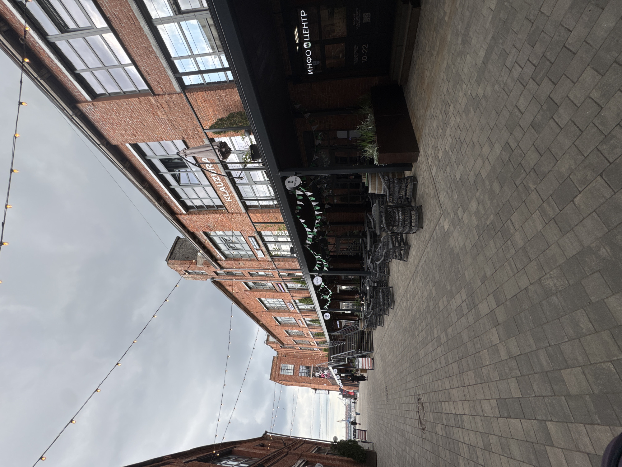 This image shows a view of a pedestrian walkway lined with brick buildings, cafes, and an information center. The walkway is paved with gray rectangular stones. Overhead, strings of decorative lights are strung across the walkway. The buildings have large windows and exposed brick facades. Several cafes have outdoor seating areas with wicker furniture. One building has a sign that reads "KLAUZURA". Another building has a sign that says "ИНФО ЦЕНТР" (INFO CENTER) in white lettering, with operating hours "10-22" below. There are a few people walking along the path. The sky is overcast, suggesting a cloudy day. The overall scene appears to be a revitalized industrial or port area, common in modern urban development in Saint Petersburg, Russia, such as the area around the New Holland Island.