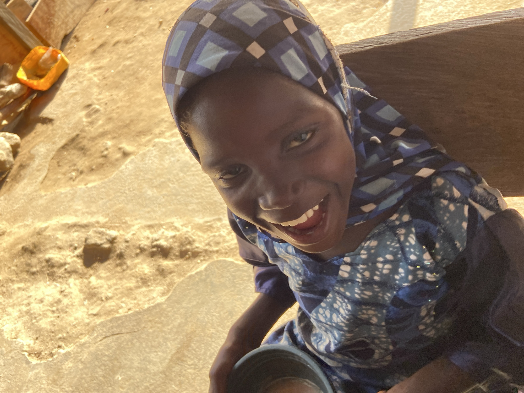 A young, dark-skinned person, possibly a child, with a broad smile and visible teeth is the primary subject of the photograph. They are wearing a blue and white patterned hijab and a blue and white patterned top. The person is holding a grey bucket or container.
The background appears to be an outdoor setting with a textured, sandy ground. To the left of the frame, there is an orange object. Behind the person, there is a wooden structure, possibly a bench or part of a building.
Given the prompt's context, it can be inferred that the location is Mararaba, Nigeria, but no specific landmarks or signs confirming the exact location are visible in the image. The environment suggests a simple, possibly rural or semi-urban setting.