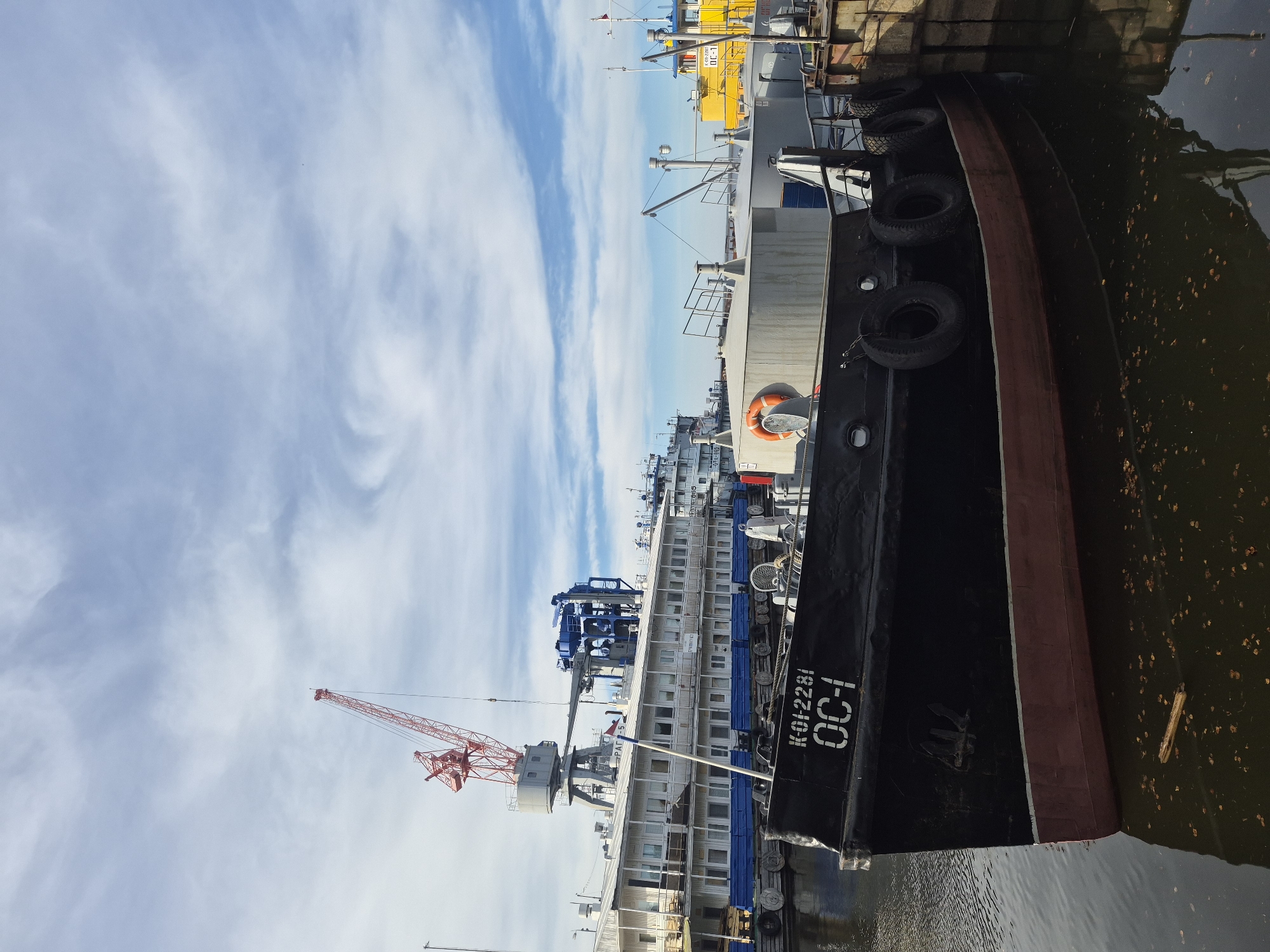 The image shows several boats docked along a waterfront in what appears to be a city. The sky is partly cloudy.
In the foreground is a section of a larger ship, painted primarily black with a red waterline. The ship is docked next to a pier and appears to have several tires along its side to act as bumpers. The ship's hull is marked with white lettering, including "K-012281" and "OC-1".
Further back are other boats and ships, some of which are white or gray with blue accents. A red crane rises above these boats. In the background, one of the boats is painted bright yellow. The water is dark and contains some debris.
