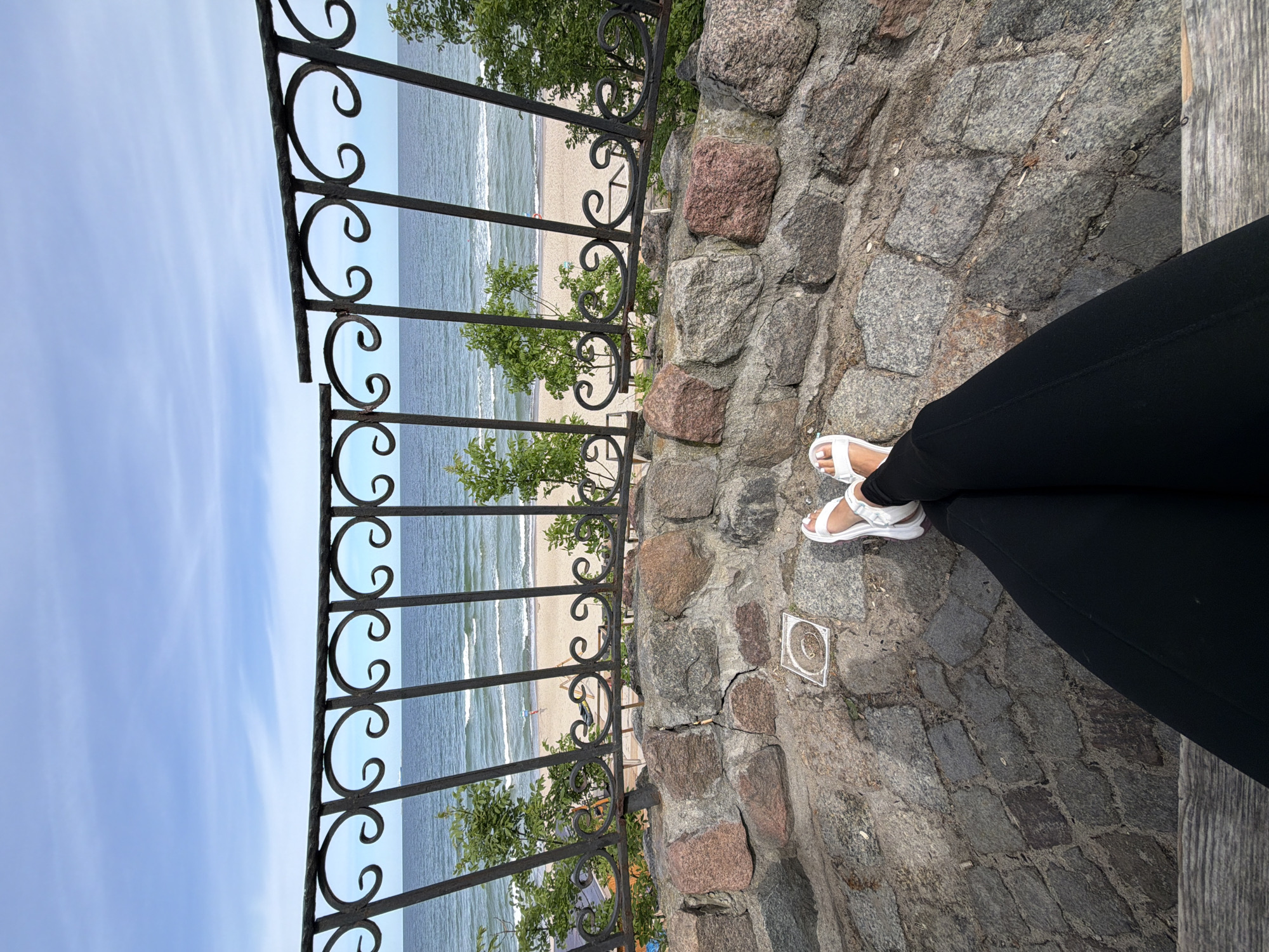 The image shows a person's legs and feet, clad in black pants and white sandals, resting on a stone-paved area. In the background, a black metal railing with decorative scrollwork overlooks a sandy beach and the sea under a bright blue sky with scattered clouds. The location is identified as being in Zelenogradsk, Russia. The scene appears to be a scenic viewpoint on a sunny day.