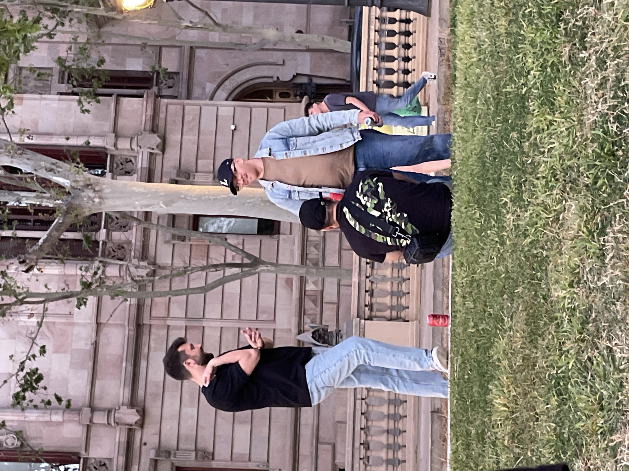 Four men stand on the street adjacent to a large stone building. A tree with bare branches partially obscures the building. The man in the foreground faces left, wearing a black shirt and light blue jeans. A red can sits on the ground near his feet. Two men stand behind him, one in a black shirt with a camouflage pattern and the other in a denim jacket over a brown shirt and a baseball cap. A fourth man stands further back, wearing a gray shirt, with his back to the camera. The setting is a grassy area next to a stone building with windows and arched doorways. The architecture of the building suggests a location in Barcelona, Spain.