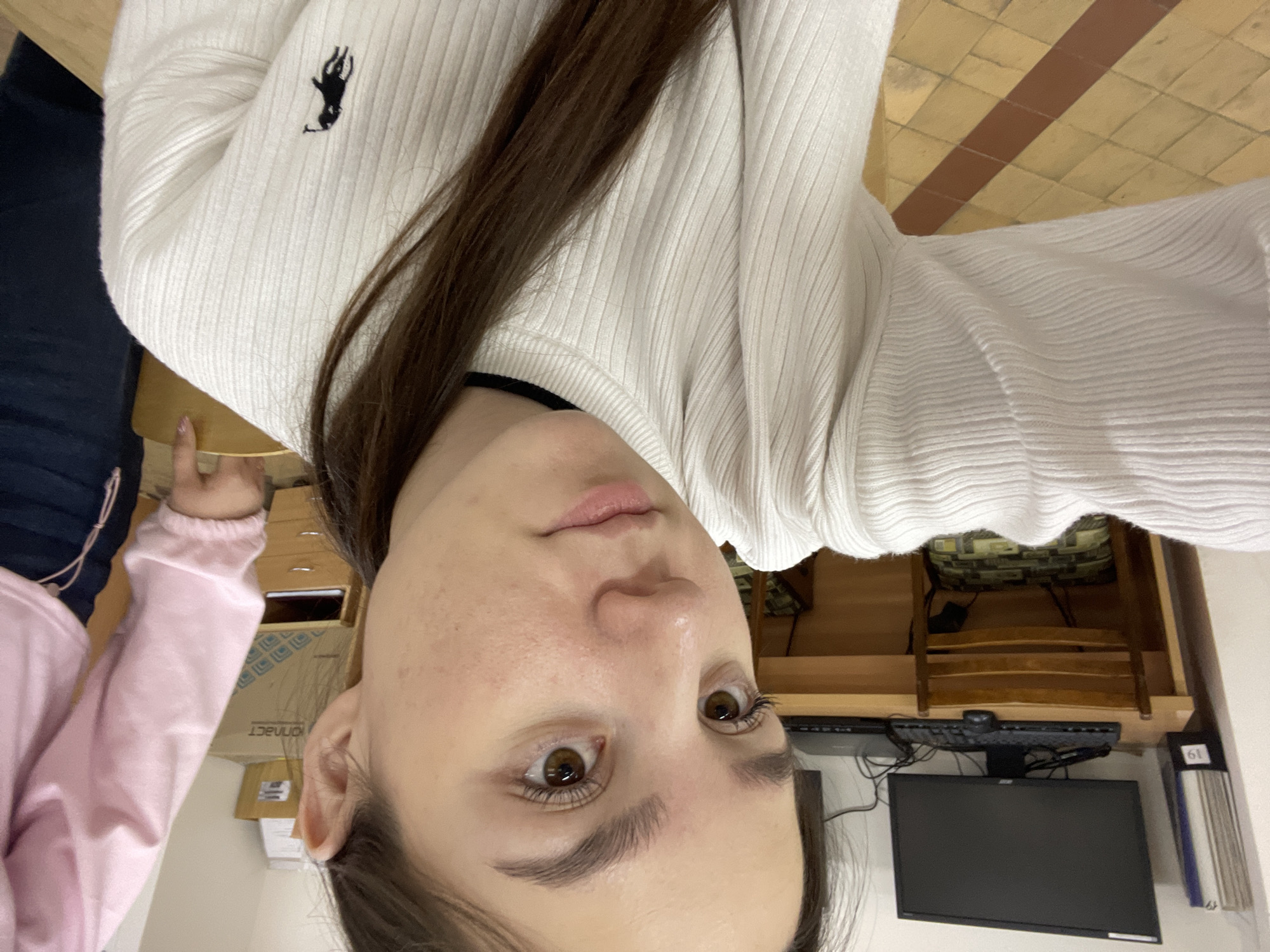 A close-up, upside-down selfie shows a woman with light skin, brown hair, and brown eyes. She is wearing a white, ribbed sweater with a small logo on the upper left side. Part of another person's arm, wearing a pink sweater, is visible in the lower left corner of the image. The background includes a computer monitor and keyboard on a desk, bookshelves, and a tiled floor. Given the presence of Cyrillic script on some items, the location is consistent with a place in Russia. There is no specific visual information to confirm the location in the city of Chaikovsky.