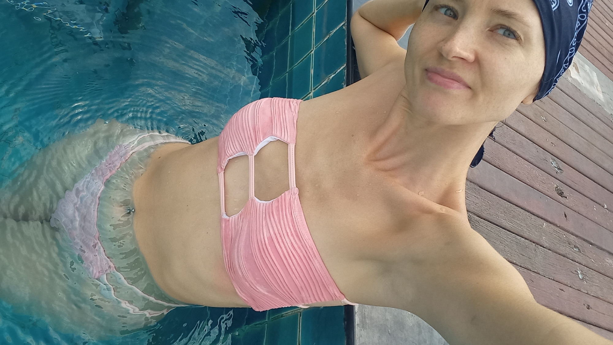 A woman wearing a pink bikini and a dark blue bandana is partially submerged in a swimming pool. She leans on a wooden surface near the edge of the pool, which is lined with dark teal tiles. The water distorts the lower half of her body. The location is Pattaya City, Thailand.