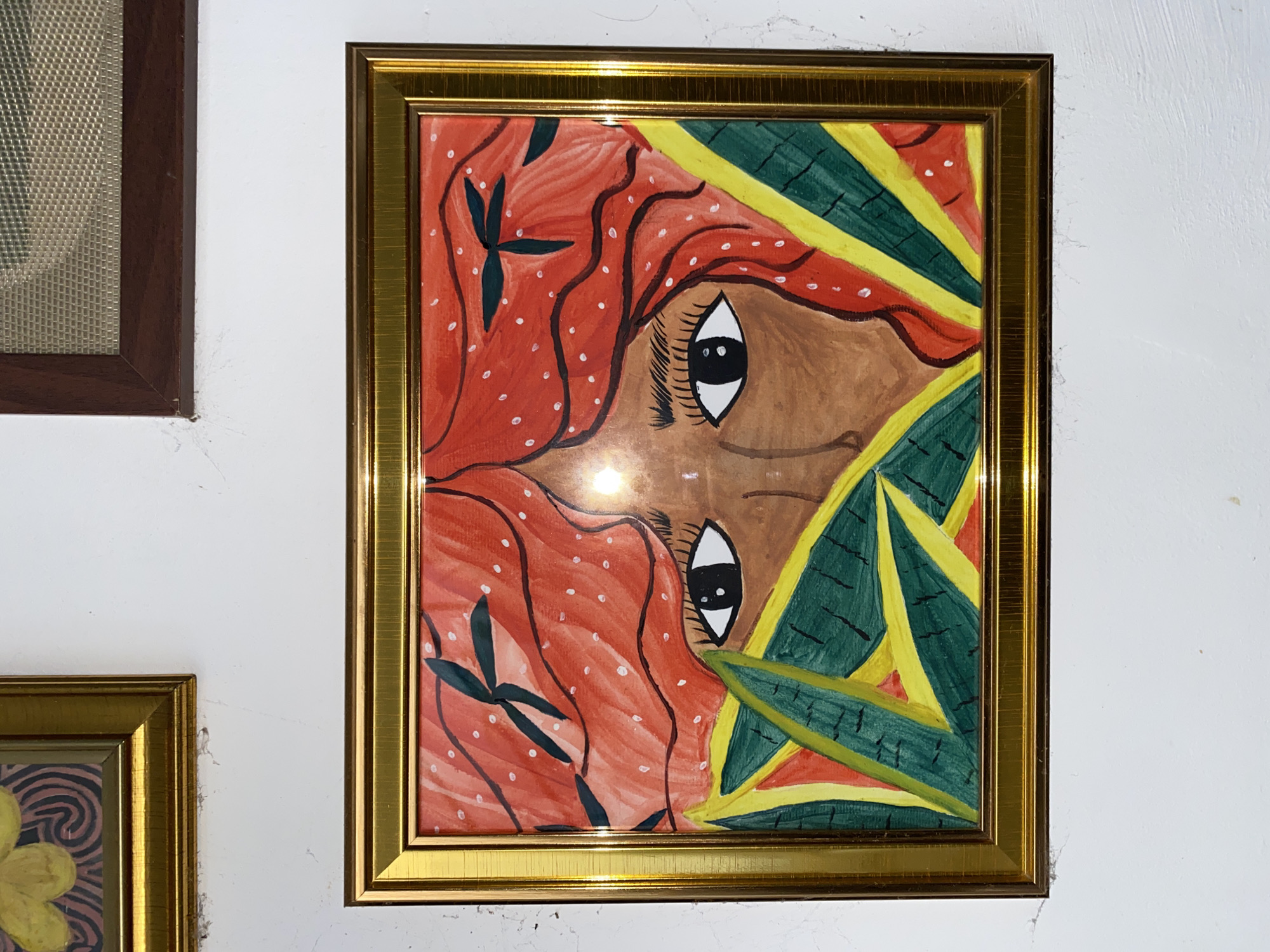 The image shows a painting of a person's face peeking through foliage. The painting is framed in gold and hangs on a white wall. Above and to the left, a portion of another framed artwork featuring a yellow flower is visible. To the upper right, a dark brown frame surrounds a textured material, possibly a woven fabric. The painting itself depicts a person with brown skin, red, wavy hair adorned with white dots, and dark eyelashes. Green and yellow leaves partially obscure the face. The background of the painting is red with white dots. The location is identified as Davao City, Philippines.