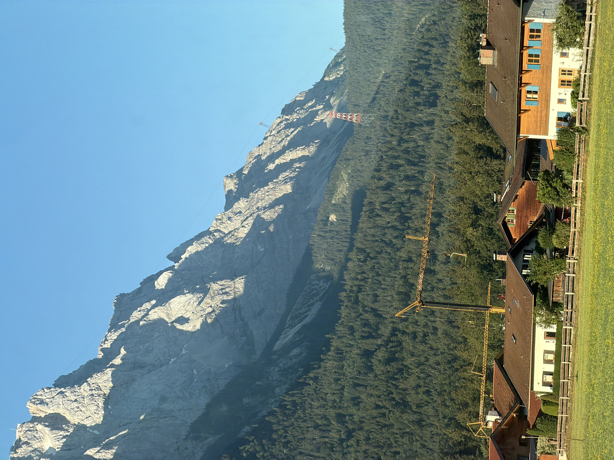 The image depicts a scenic view in Grainau, Germany, with a towering, rugged mountain dominating the upper portion of the frame under a clear blue sky. The mountain face is stark and rocky, with visible shadows indicating the direction of sunlight. Below the mountain, a dense, dark green forest stretches across the mid-ground, forming a lush backdrop.
In the foreground, a cluster of traditional Bavarian-style houses with brown tiled roofs and wooden accents are visible. One house in particular stands out with white walls and bright blue shutters on its windows. A wooden fence runs along the edge of a vibrant green meadow, dotted with yellow wildflowers, separating the houses from the natural landscape.
Adding a contrasting element to the natural beauty, several construction cranes are present in the mid-ground, near the forest line and above the houses, suggesting ongoing development. Higher up on the mountain slopes, two red and white transmission towers are visible, indicating infrastructure related to power lines or possibly a ski lift system. The scene is one of natural grandeur juxtaposed with human habitation and construction.