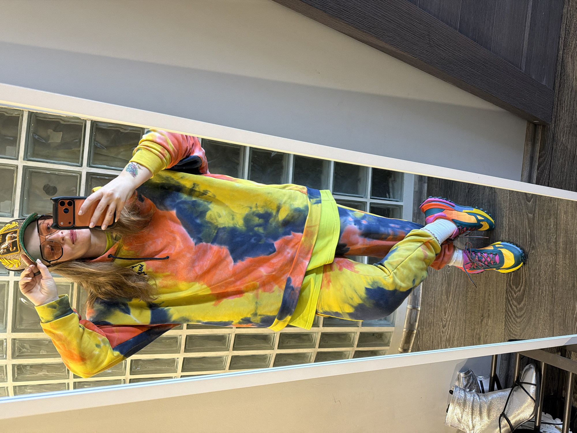 A woman takes a selfie in a mirror. She wears a tie-dye tracksuit in yellow, orange, and navy blue, and bright running shoes. She also wears an olive green cap with a decorative patch, and orange-tinted eyeglasses. Her right arm has a small tattoo on the wrist. Her left hand holds the cap. Behind her in the mirror are glass blocks, and the wood-tone wall and doorframe. Below, near the floor, are a pair of silver boots with black ties. The location in the city of Moscow, Russia cannot be determined.