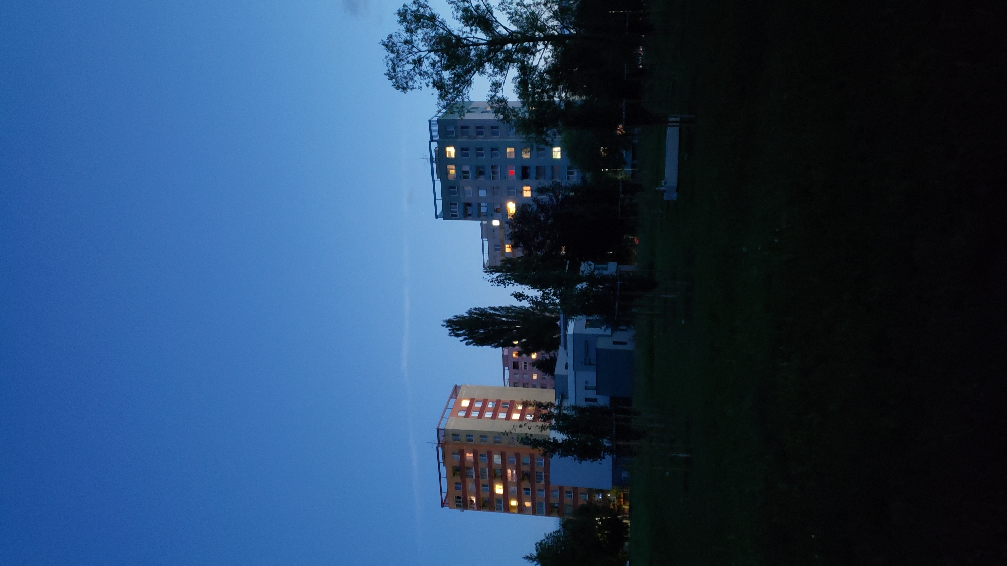 The image shows a residential area at dusk in Prešov, Slovakia. Several multi-story apartment buildings are visible, with lights on in some of the windows, indicating that it is evening. The sky is a deep blue, suggesting the transition from day to night. In the foreground, there is a dark grassy area with some young trees planted in rows. The overall scene depicts a typical urban landscape during the twilight hours.