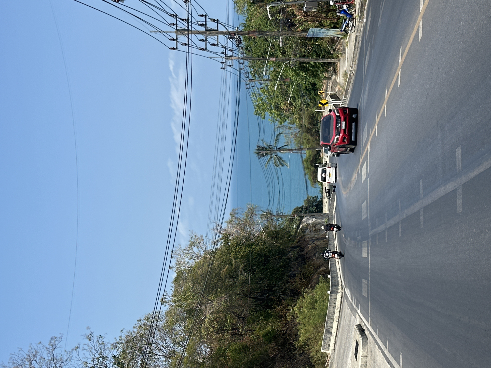 The image shows a coastal road in Baan Chaweng Noi, Thailand, under a clear blue sky. The road runs alongside the ocean, visible beyond trees and power lines. A red car and a white car are on the road, followed by two motorcycles. Lush green vegetation covers the area bordering the road.