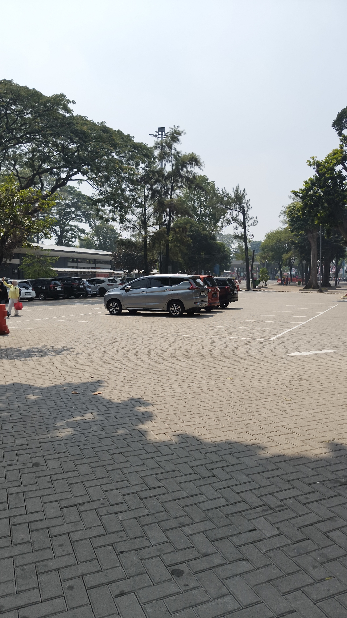 This is a full-shot, eye-level, vertical photograph of an outdoor parking lot in South Jakarta, Indonesia. The parking lot is paved with interlocking brick pavers and is marked with white lines to designate parking spaces. The background features a row of trees and a low-rise building. Several cars are parked in the lot, including a silver SUV in the foreground. One person, wearing a yellow jacket and holding a red bag, is standing on the left side of the frame. The sky is clear and bright.