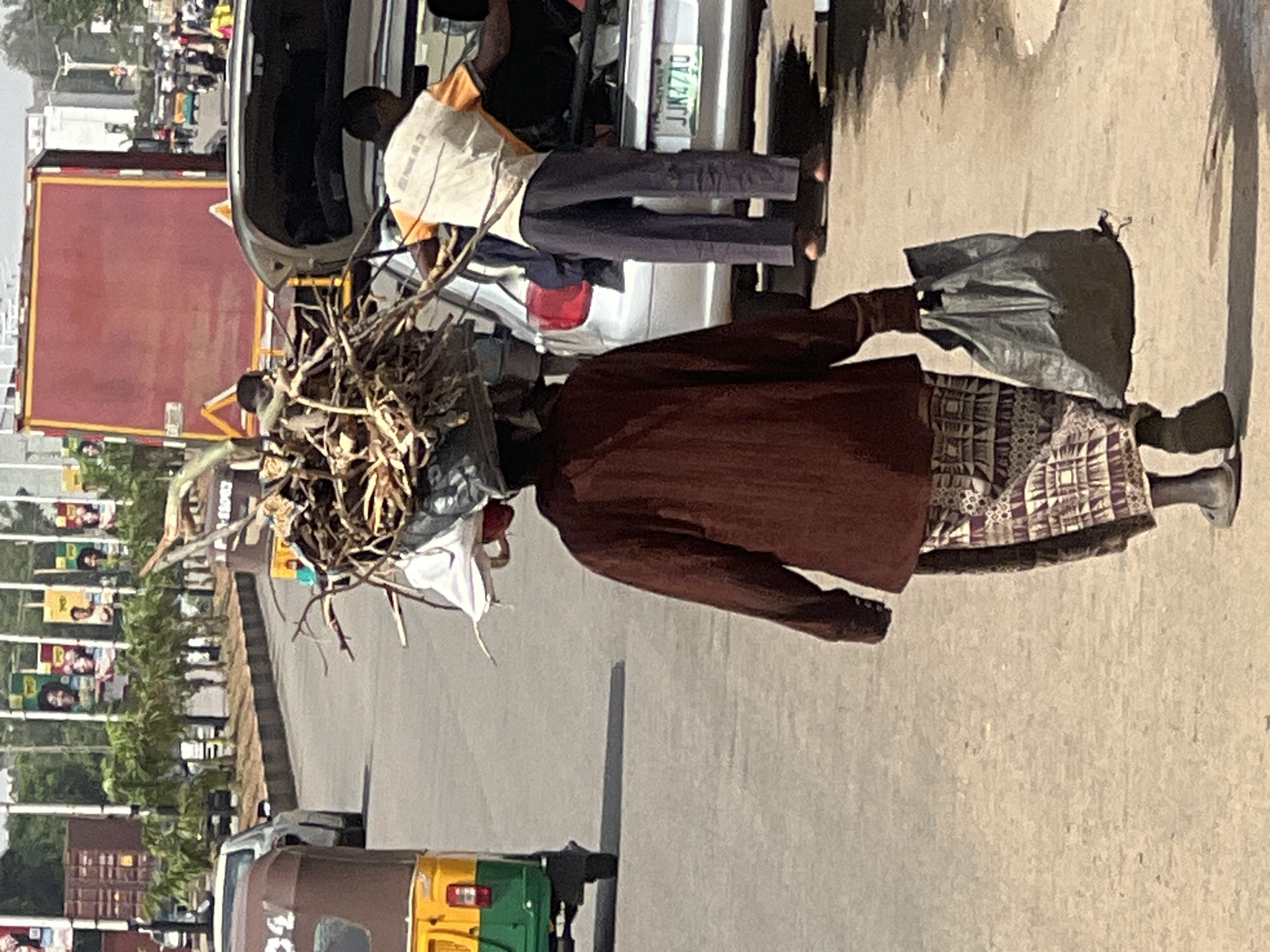 A person carries a large bundle of branches on their head while walking on a paved street in Barakin Lerawi, Nigeria. The person wears a dark red shirt, patterned skirt, and gray shoes. They carry a gray bag in their left hand. Behind them is a silver car with license plate JJN27AD. Further back, there is a red truck and a yellow and green auto rickshaw. Advertisement posters line the street.