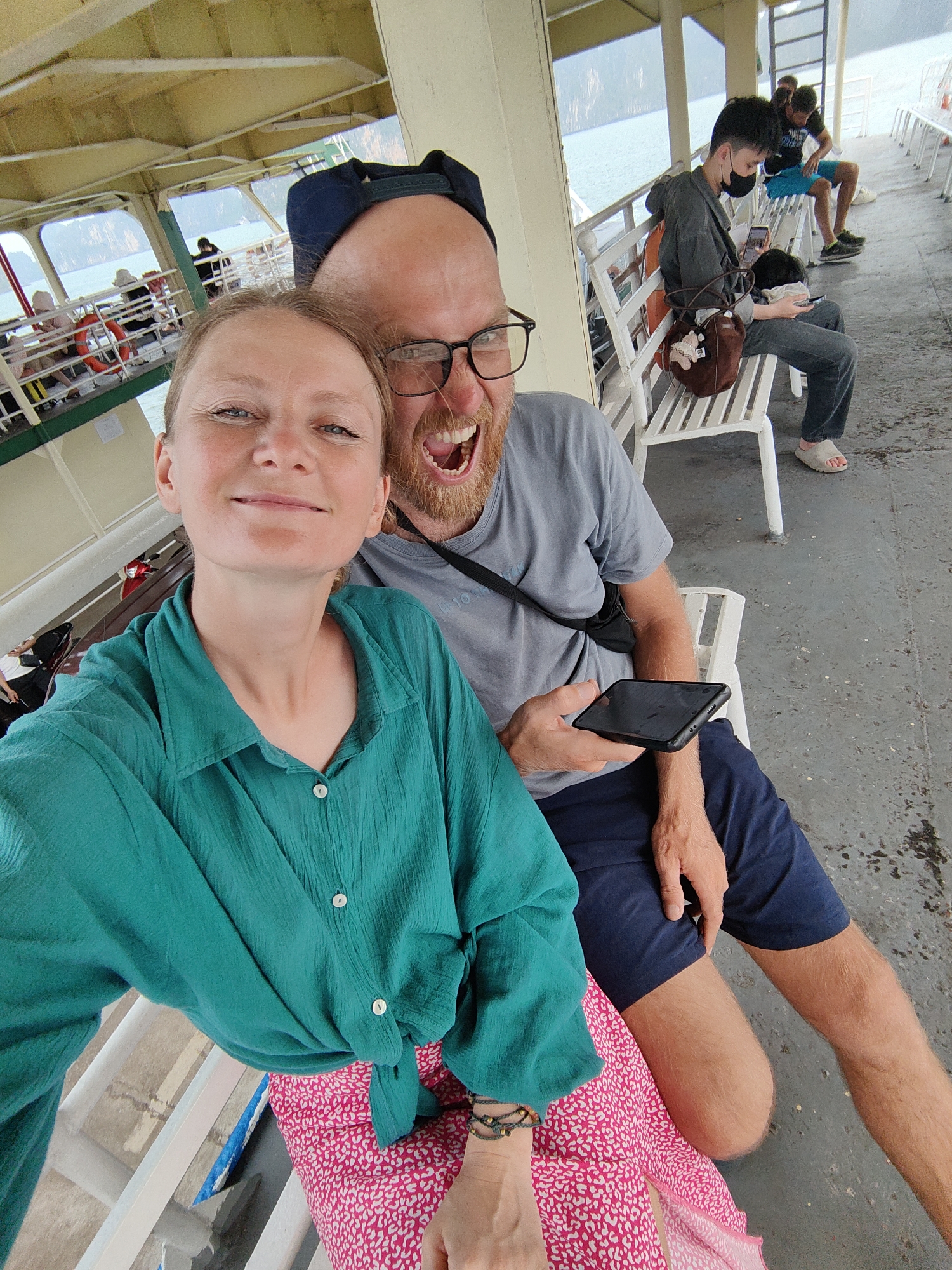 A selfie captures a fair-skinned woman with a serene smile, donning a green button-up shirt tied at the waist over a pink-patterned skirt, seated on a white slatted bench. Beside her, a fair-skinned man with a red beard and glasses, wearing a blue baseball cap and a gray t-shirt, holds a smartphone while smiling widely with his mouth open. They are both sitting on the bench on what appears to be a ferry, with other passengers visible on adjacent benches. One passenger, wearing a mask, is engrossed in a phone. The backdrop reveals a body of water surrounded by elevated terrain. The location is identified as Tuan Chau Ward, Vietnam.