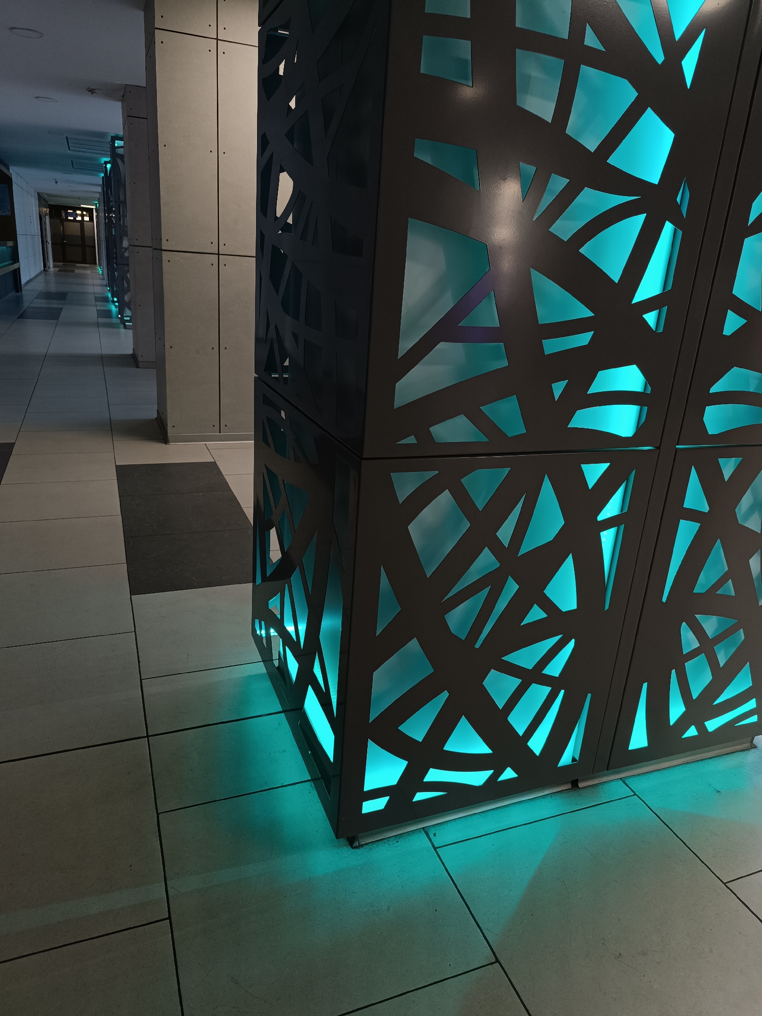 The image depicts an indoor scene in Sofia, Bulgaria. A corridor with tiled flooring is visible. In the foreground is a decorative column-like structure composed of dark gray panels with irregularly shaped cutouts, illuminated from within by a turquoise light. This illumination creates a striking contrast against the dark gray of the panels and casts a glow onto the surrounding floor tiles. In the background, a series of similar columns or structures are visible, extending along the corridor. A door can be seen at the end of the corridor. There are no people present in the image.