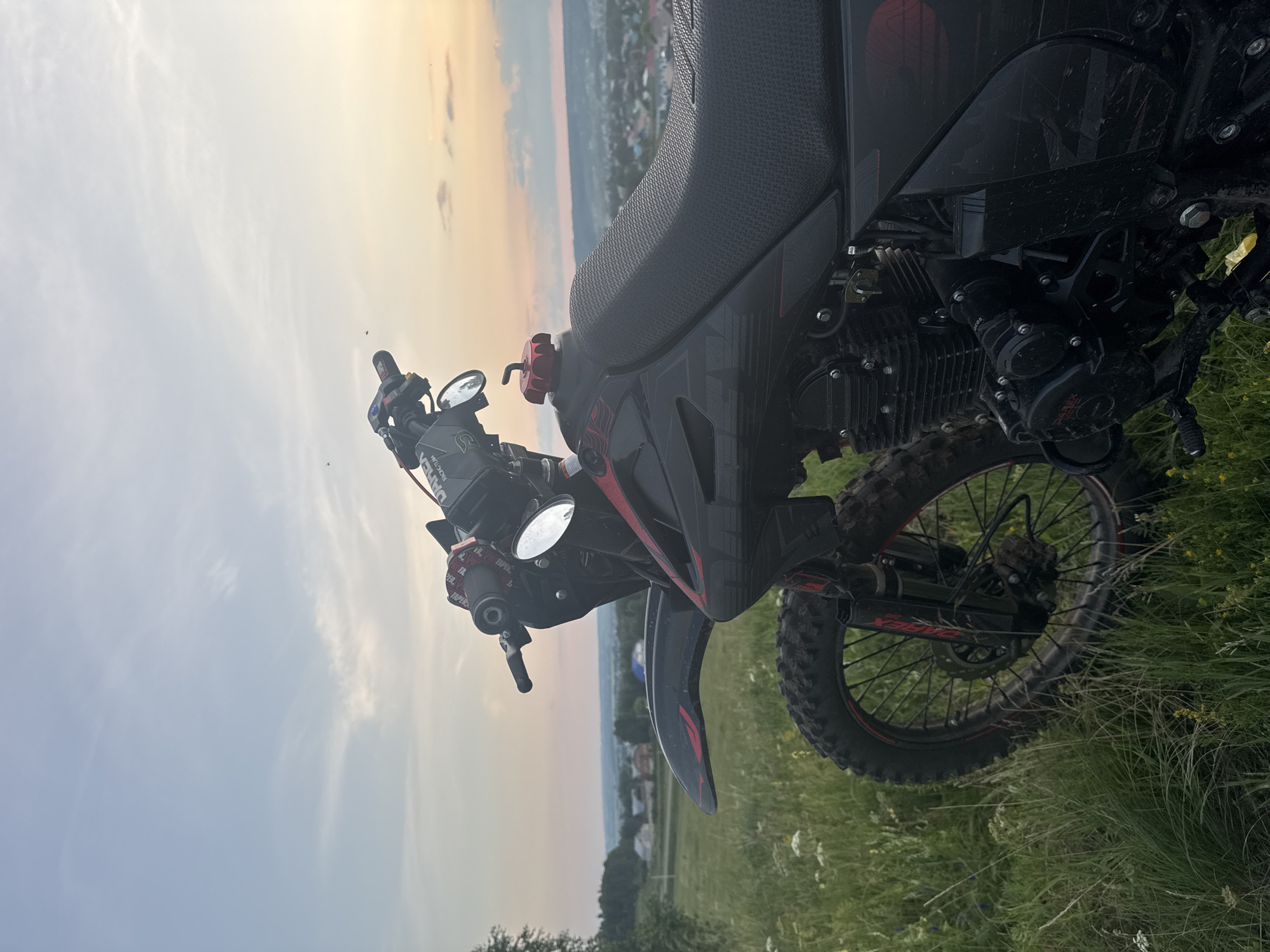 The image shows a dirt bike parked in a grassy field at sunset, with a village and hills visible in the background. The sky is painted with warm hues of orange and yellow, indicative of the setting sun. The dirt bike is black with red accents and features knobby tires, suggesting it's designed for off-road use. The handlebars, mirrors, and a portion of the seat are visible. The rear wheel and part of the engine are also in focus. The grass around the bike appears tall and somewhat overgrown, with some wildflowers interspersed. The scene evokes a sense of peacefulness and adventure, perhaps after a day of riding. The location is identified as Sharan, Russia. The time of day is sunset. There is no visible text in the image.