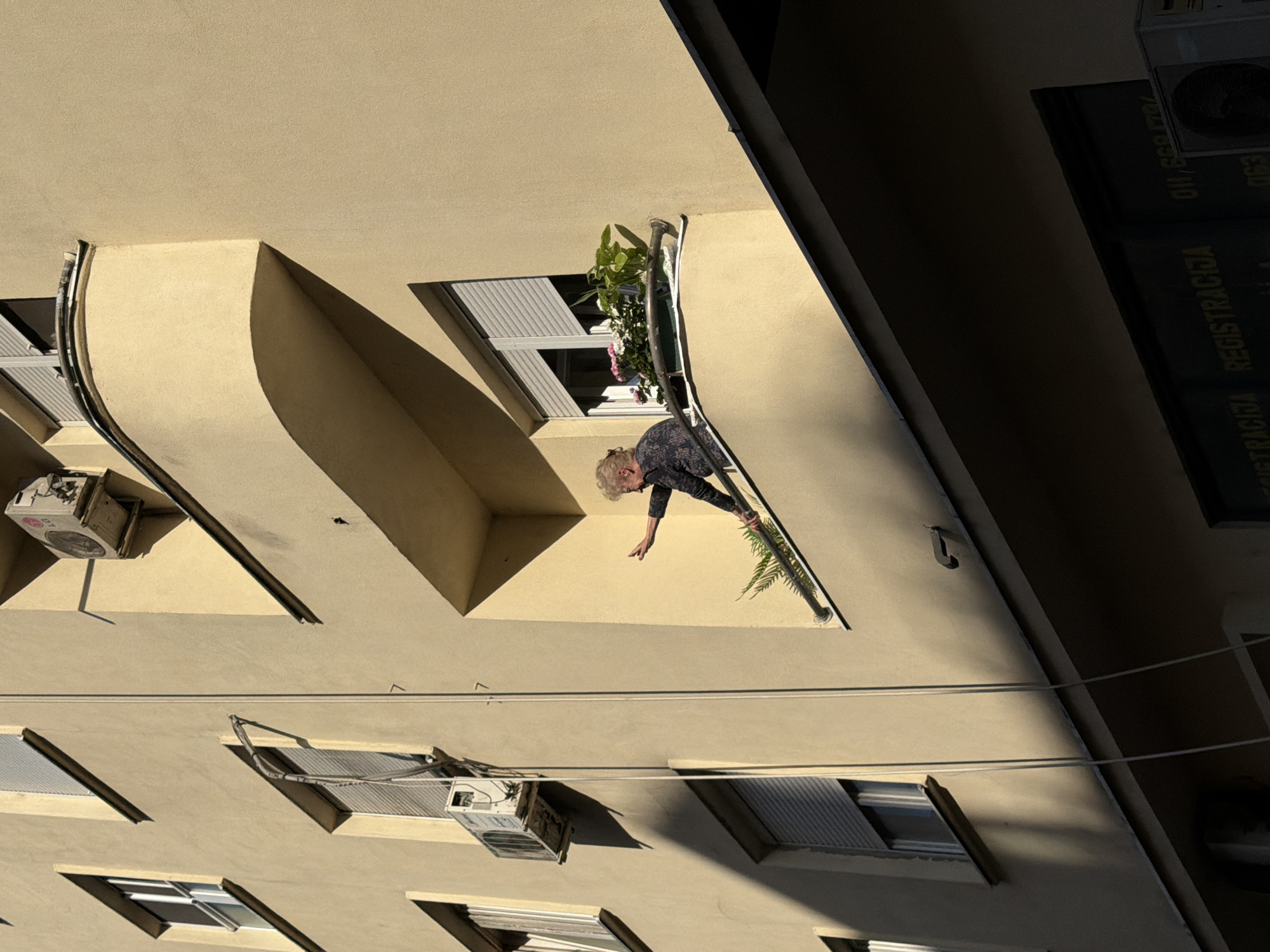 The image depicts an elderly woman leaning over a balcony in Belgrade, Serbia. She is reaching down towards the street below. The balcony has a flower box with plants. The building's facade is a light yellow color. There are windows with louvered blinds and air conditioning units attached to the building. Utility wires run across the building's face.