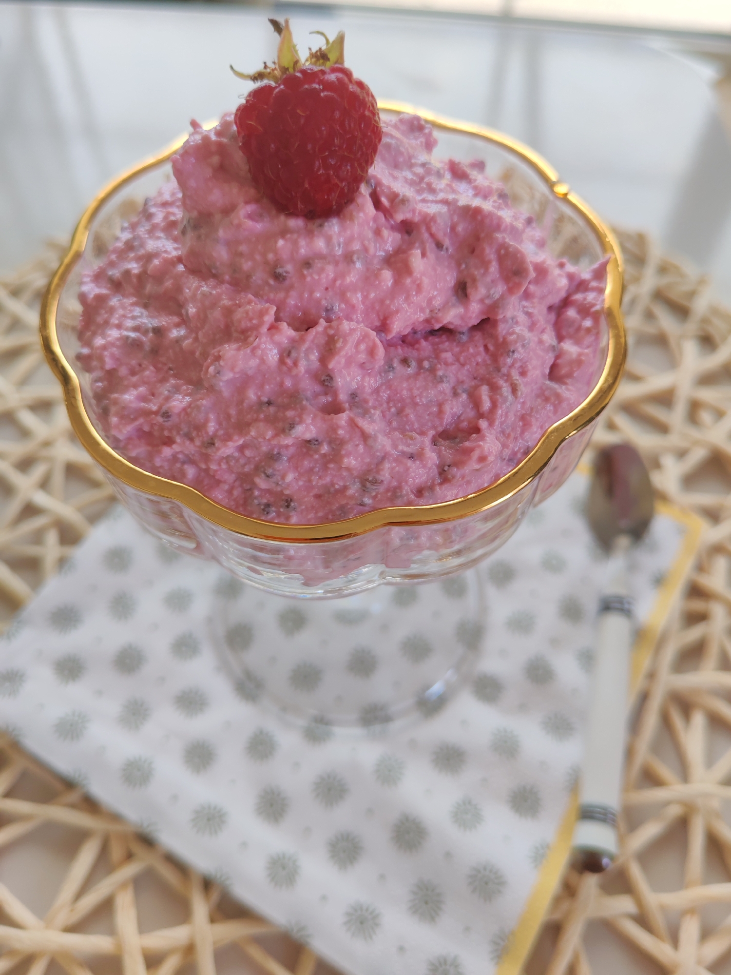 A close-up, eye-level shot shows a glass dessert bowl filled with a pink, creamy mixture, possibly yogurt or pudding, with visible seeds and berry pieces. A single, ripe raspberry with its stem and leaves rests on top of the mixture. The bowl has a decorative gold rim and sits on a woven placemat. A napkin with a subtle floral pattern is placed beneath the bowl, and a portion of a spoon with a decorative handle is visible to the right. The background is blurred, suggesting an indoor setting with natural light. The scene does not provide any information that can be used to determine its location in Sabunchu Raion, Azerbaijan.