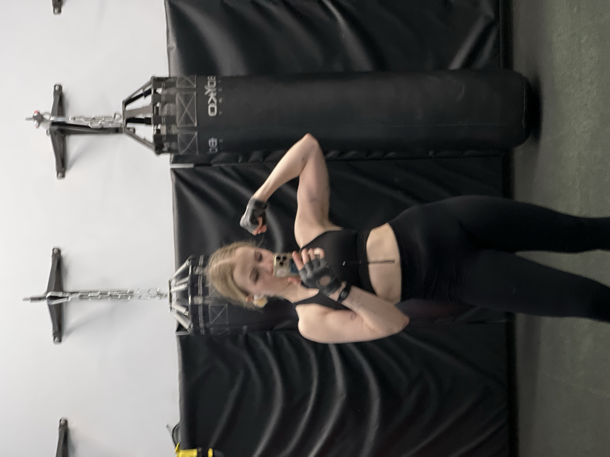 A woman in black workout attire poses in front of a boxing bag in what appears to be a gym or workout space. The background includes padded black walls. A boxing bag is suspended from a wall-mounted bracket. The woman is taking a selfie with a mobile phone, flexing one arm. The location cannot be reliably determined from the image, and the city of Khimki, Russia is not apparent.