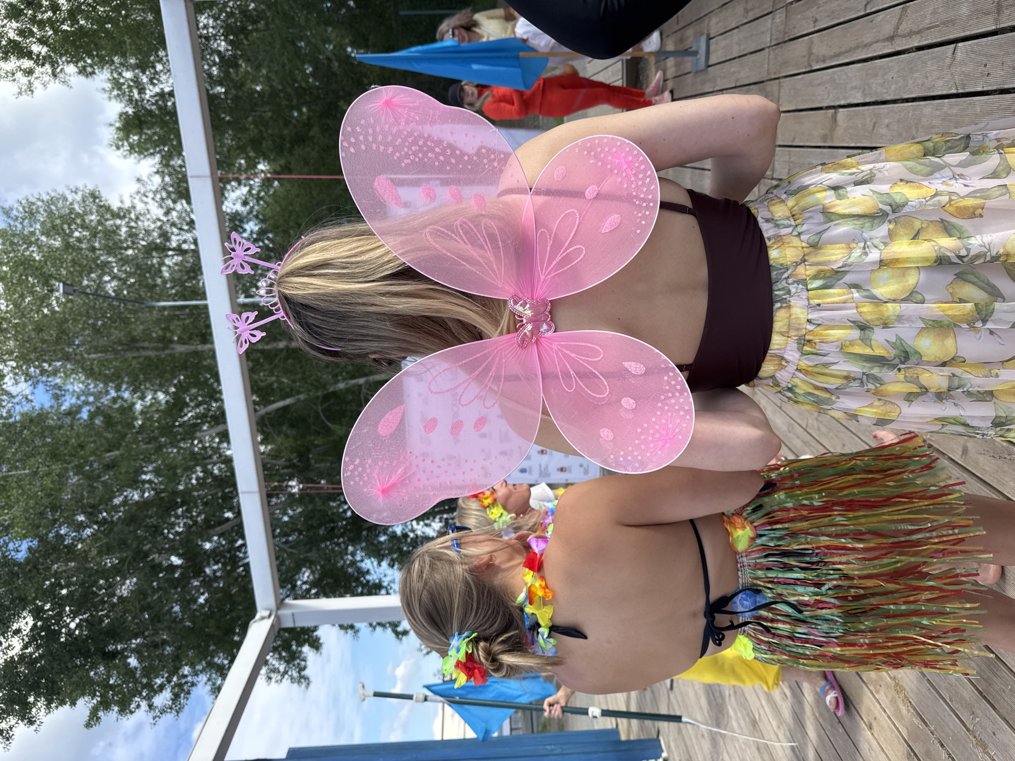 Two women are viewed from behind at an outdoor event. The woman on the left wears a black bikini top, a colorful Hawaiian lei, and a fringed grass skirt. She has a flower in her hair. The woman on the right wears a maroon camisole and a sheer, pleated skirt with a lemon pattern. She has large pink butterfly wings attached to her back and a pink tiara with butterflies on her head. In the background, other people, flags, and trees are visible. The event appears to be taking place on a wooden deck, possibly near water. The sky is cloudy. The location is identified as Chaikovsky, Russia.