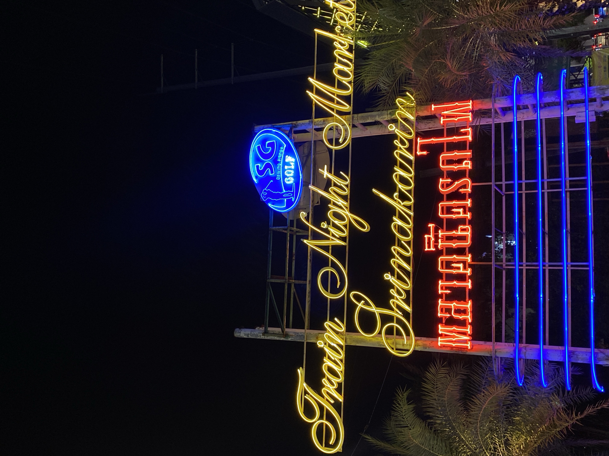 The image shows a brightly lit sign that reads "Train Night Market Srinakarin" in Bangkok, Thailand. The words "Train Night Market" are in large, cursive yellow neon lettering, while "Srinakarin" is in a similar, but slightly smaller yellow neon script. The words "Night Market" are vertically stacked. To the right, vertically stacked and in red neon, are the letters "NIGHT MARKET". Behind the sign, there is a series of blue neon vertical poles or tubes. To the left is a circular blue sign with "SG Golf" written in white, along with an image of a golfer. The sign is on a metallic structure. Palm trees are visible at the base and top of the sign. The background is dark.