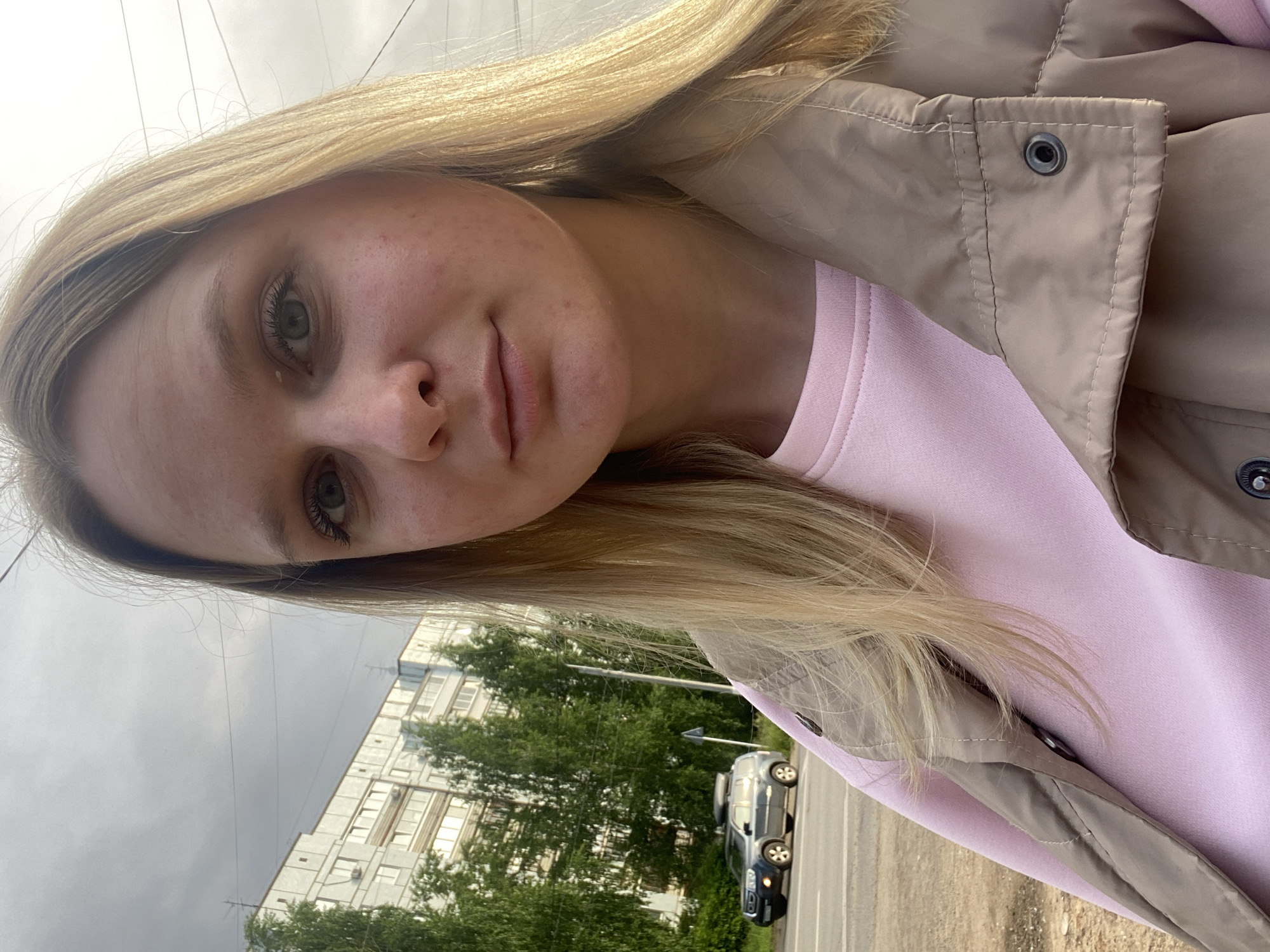 A young, fair-skinned woman with blond hair and green eyes is taking a selfie. She is wearing a light pink sweatshirt and a beige jacket. Behind her, a multistory apartment building with white windows is visible, partially obscured by green trees. A gray SUV is parked on the roadside in front of the trees. The sky is overcast. The image appears to be taken in an urban environment, likely in the city of Chaikovsky, Russia, based on the location provided.