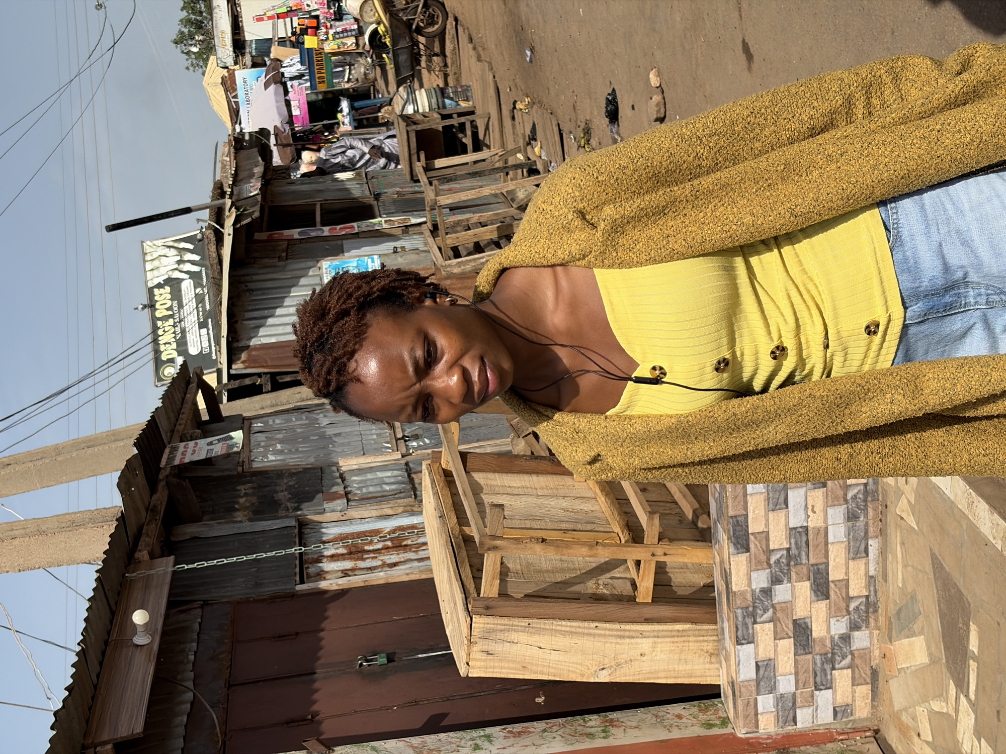 The image captures a woman with short, curly brown hair, wearing a yellow top, a mustard-colored cardigan, and light blue jeans. She has earphones in and is positioned in front of a building constructed of corrugated metal. The building appears to be a commercial space, with signage visible, including "DEAGE POSE." The environment suggests a bustling, urban area, likely in Gura Top, Nigeria, given the prompt. The scene includes various makeshift structures and stalls that are typical of the area.