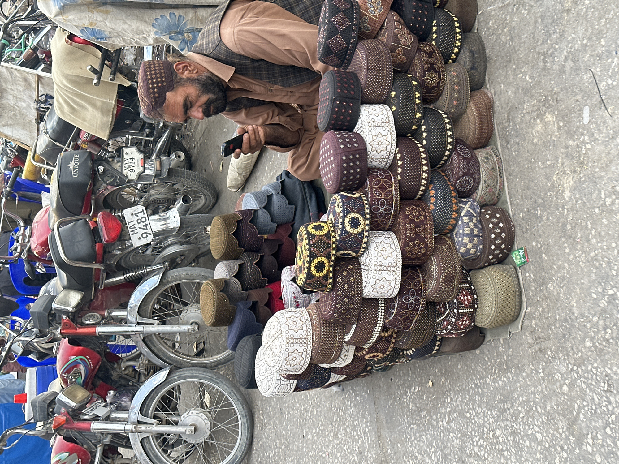 In Quetta City Tehsil, Pakistan, a man sits on the ground surrounded by a large display of embroidered caps. He is wearing a brown shirt and a patterned vest, and he is looking at his phone. The caps are stacked in pyramids and piles, featuring intricate embroidery in various colors and patterns, with the majority being shades of brown, white, and maroon. Behind the caps, several red motorcycles are parked, suggesting a market or roadside stall environment. One motorcycle has a license plate that reads "MAT 9481", and another "MAN 9714". The ground is paved and appears worn, with some debris scattered about. The overall scene depicts a bustling marketplace setting.