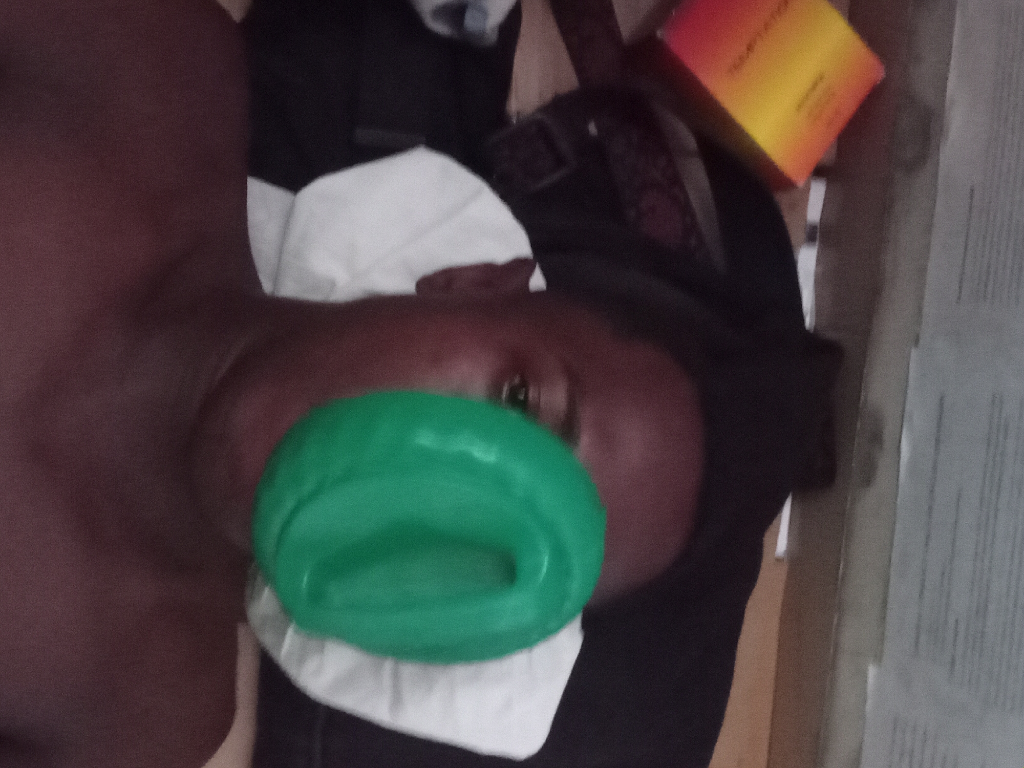 A close-up, somewhat blurry photo shows a person's face partially obscured by a green, donut-shaped object. The person appears to be lying down, as their head is tilted to the side. A white cloth or pillow is visible beneath their head, and various objects are scattered around, including a box with orange and yellow tones, and some dark clothing. The location and specific details about the city of Akwete, Nigeria are not discernible from this image.