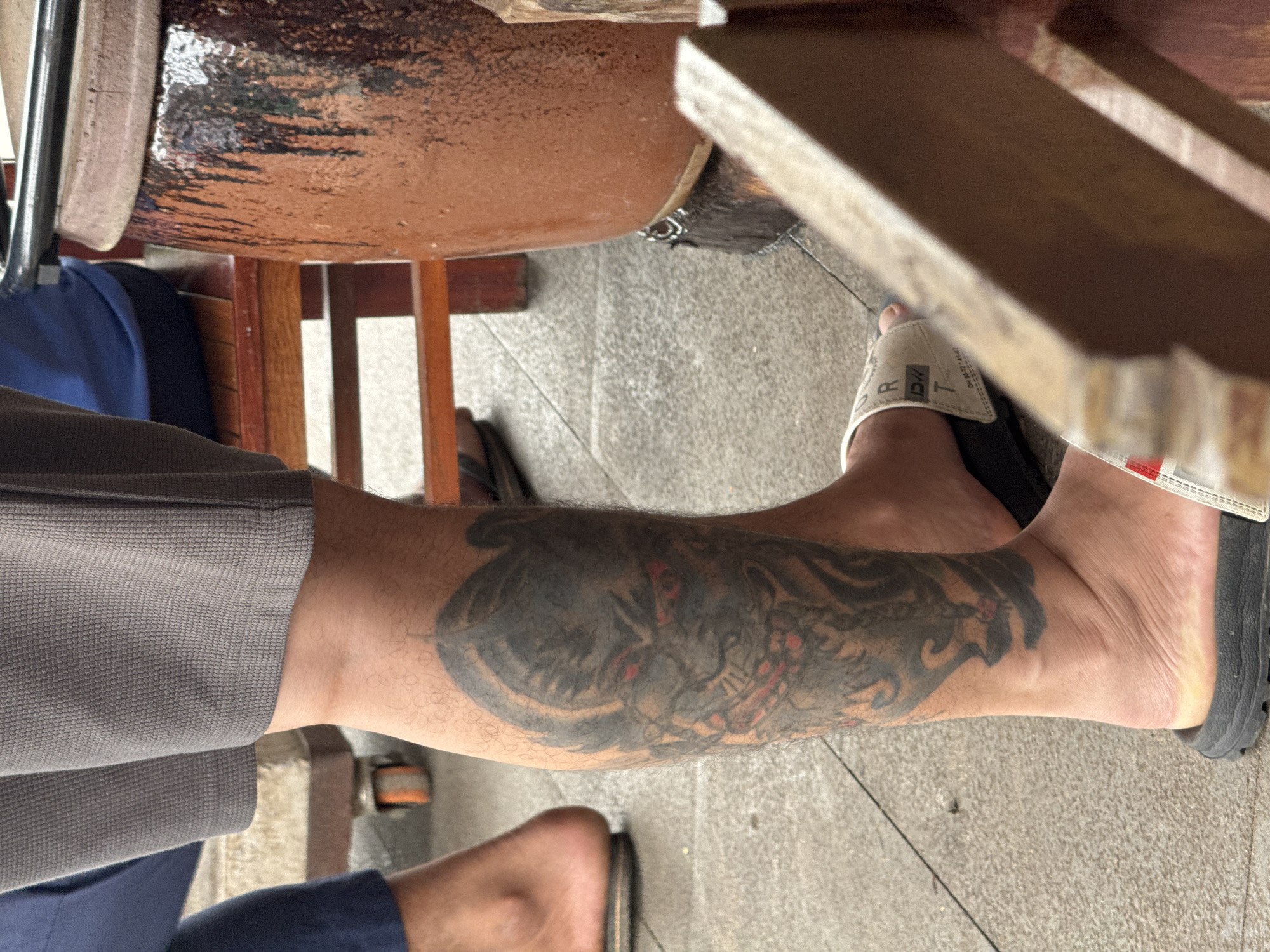 In Siem Reap, Cambodia, a close-up shot focuses on a person's lower legs and feet. They are wearing grey shorts and flip-flops. A large, detailed tattoo of a creature's face, possibly a demon or monster with red eyes, covers the calf of the left leg. The tattoo is predominantly in black and grey tones, with some red accents in the eyes and mouth. The background shows a textured brown ceramic pot with a dark, drippy glaze, and parts of a wooden structure, suggesting an outdoor or semi-outdoor setting. The ground appears to be paved with light-colored tiles.