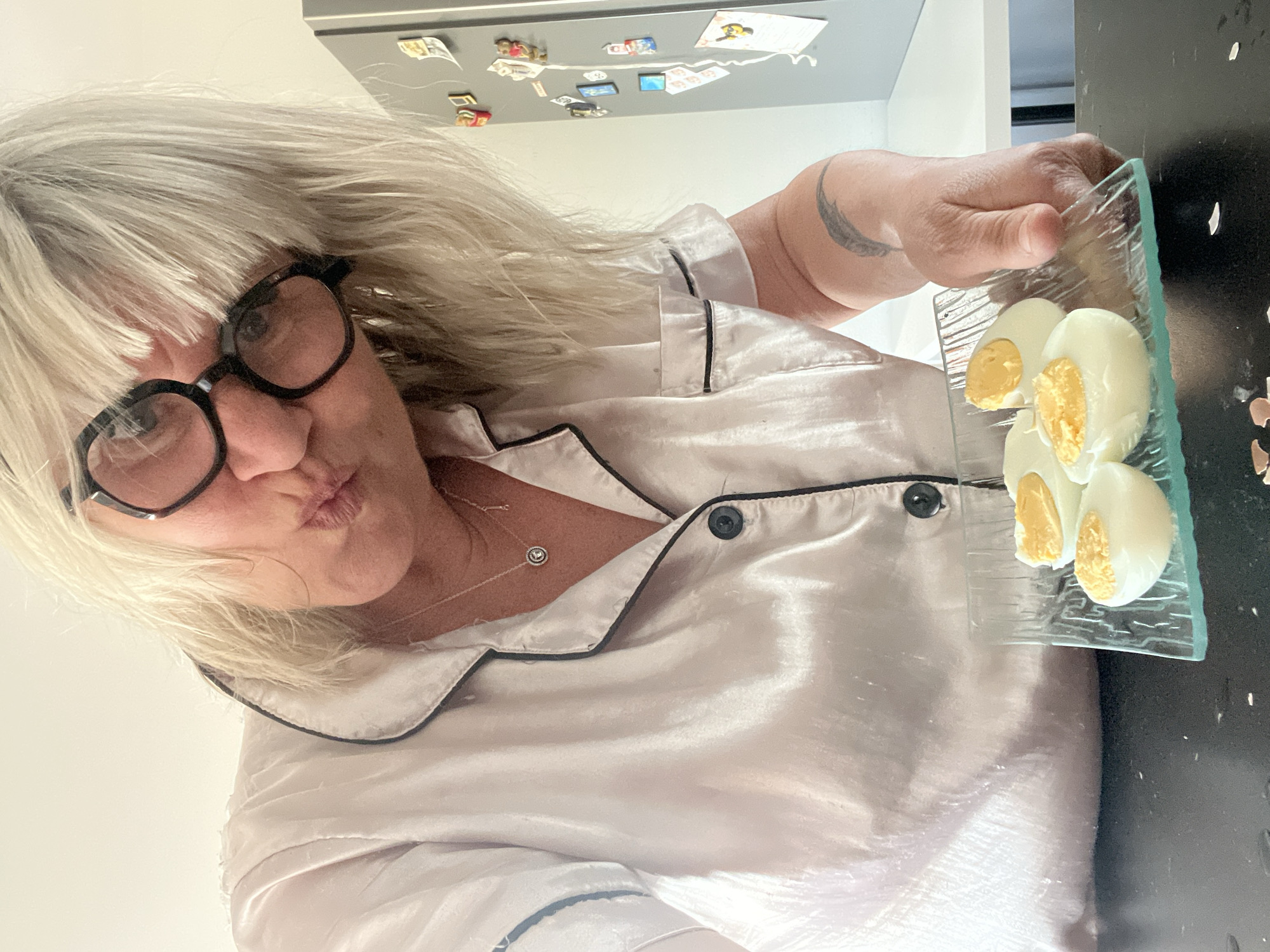 A woman with blonde hair and glasses is holding a plate with four halved hard-boiled eggs. She is wearing a satin pajama top with black piping. The background includes a refrigerator with magnets and a dark countertop. The location is Chambœuf, France.