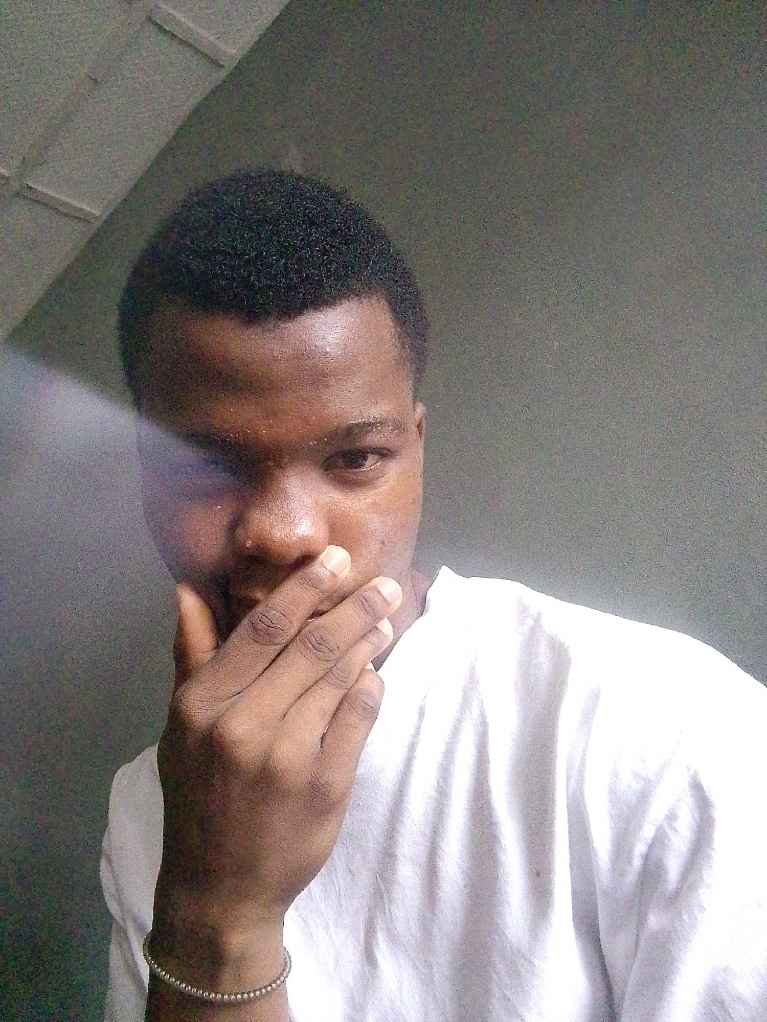 A medium, eye-level shot shows a person, likely male, with dark skin and short, dark hair. The person is wearing a white shirt and has a bracelet on their left wrist. Their left hand is raised, with the fingers partially covering the lower part of the face, near the mouth. The person is looking directly at the camera, though the left side of the face is obscured by a light or shadow. The background appears to be a gray wall, possibly indoors. The scene's location is given as the city of Unknown, Unknown.