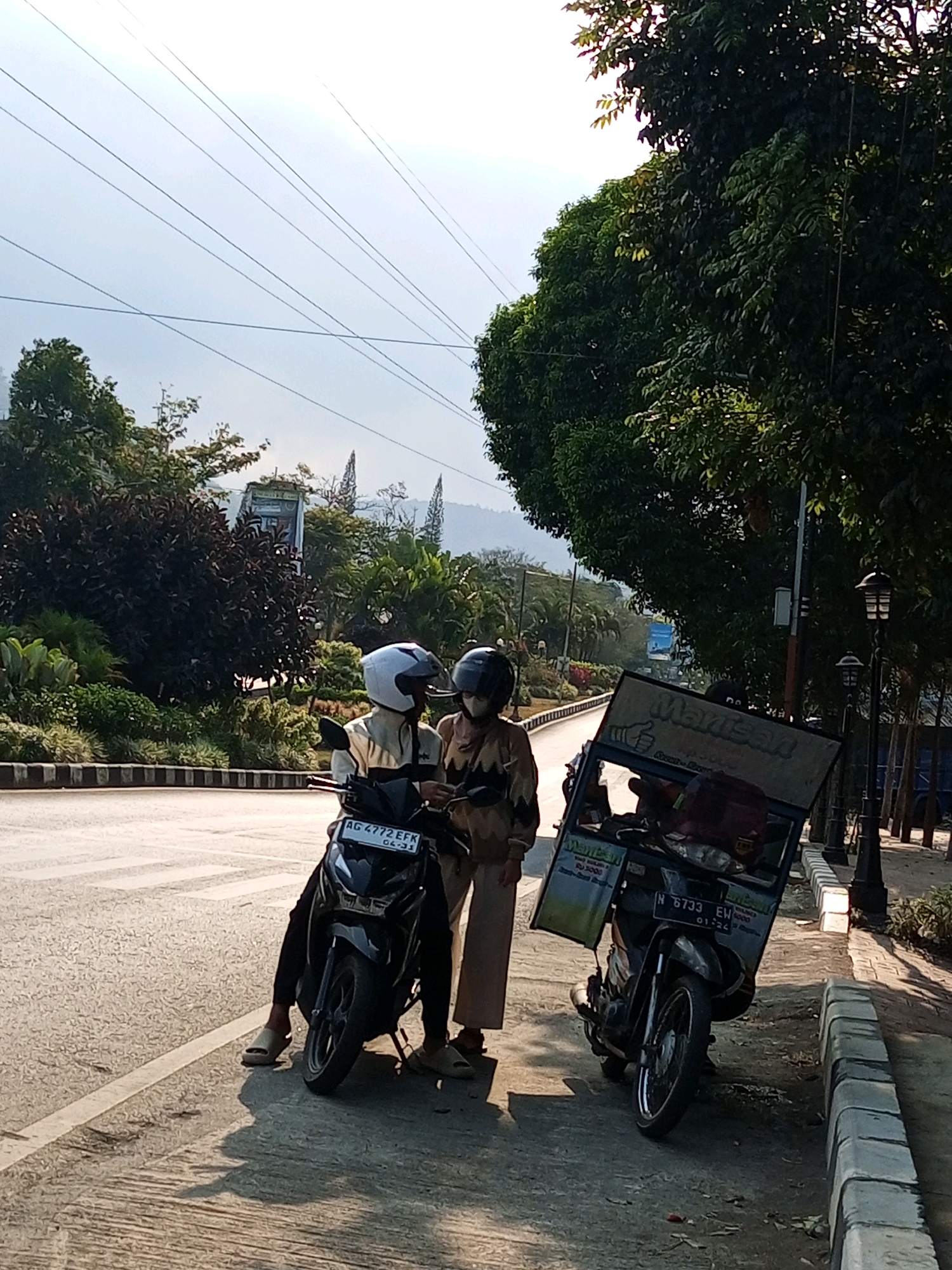 In Batu, Indonesia, two individuals wearing helmets stand next to two motorcycles on the side of a road. The motorcycle on the left, a black scooter, has a license plate reading "AG 4772 EFK 04-21". The rider of this scooter, wearing a light-colored jacket and white helmet, appears to be looking at a phone held by the second individual. The second individual, wearing a light brown hooded garment and a dark helmet, is standing to the right of the scooter and is holding the phone. To the right of these two is a second motorcycle, also a scooter, with a large rectangular box attached to its rear, suggesting it's used for delivery. This motorcycle has a license plate reading "N 6733 EW 01-24". The background features lush green trees, foliage, and a clear sky, indicating a sunny day. A paved road with a pedestrian walkway and a concrete curb is visible. In the distance, mountains and some structures are discernible.