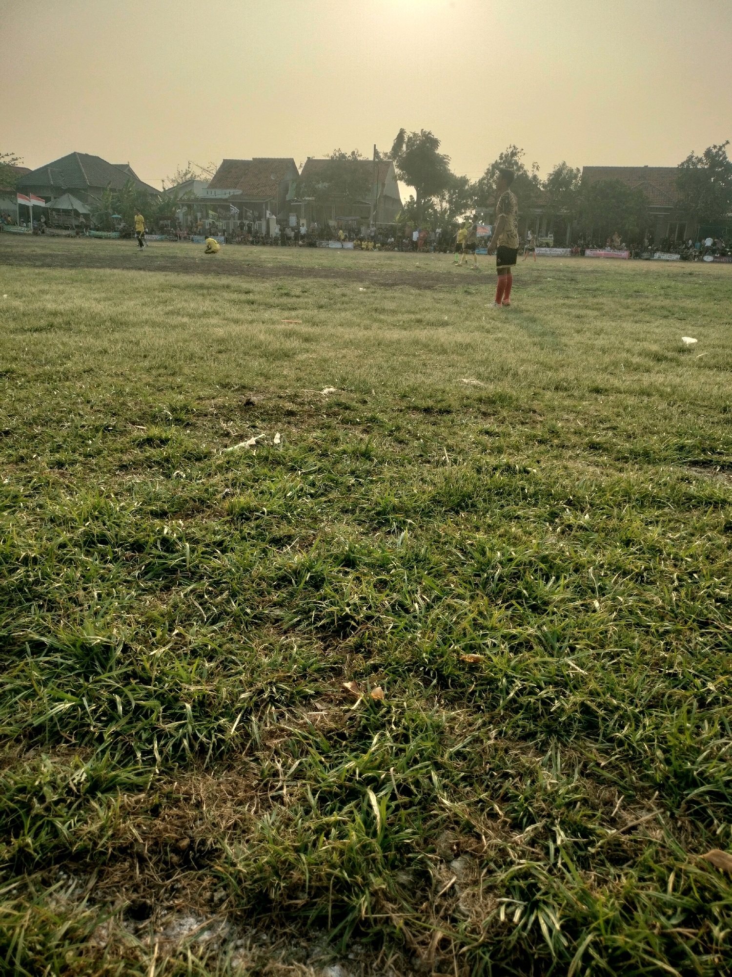 In Kalisari, Indonesia, a wide, uneven grassy field serves as the setting for a soccer match. The field is largely covered in green grass, but patches of dry, brown earth and debris are scattered throughout, indicating a lack of consistent maintenance. In the background, a line of houses with reddish-brown roofs forms the boundary of the field, with trees and foliage interspersed between them. A hazy, yellow-orange sky suggests either a sunrise or sunset, casting a diffused light over the scene.
Several individuals are present on the field. In the mid-ground, a young person wearing a yellow and brown patterned shirt and red socks stands facing away from the camera, appearing to be a player. To their left, another individual in a yellow jersey is crouching low on the field. Further back, numerous spectators are gathered behind a makeshift barrier, watching the game. A few people are also visible in the distance, scattered across the field. Two Indonesian flags are displayed prominently on the left side of the background. The overall impression is of a casual, local soccer game taking place in a rural or semi-rural area.